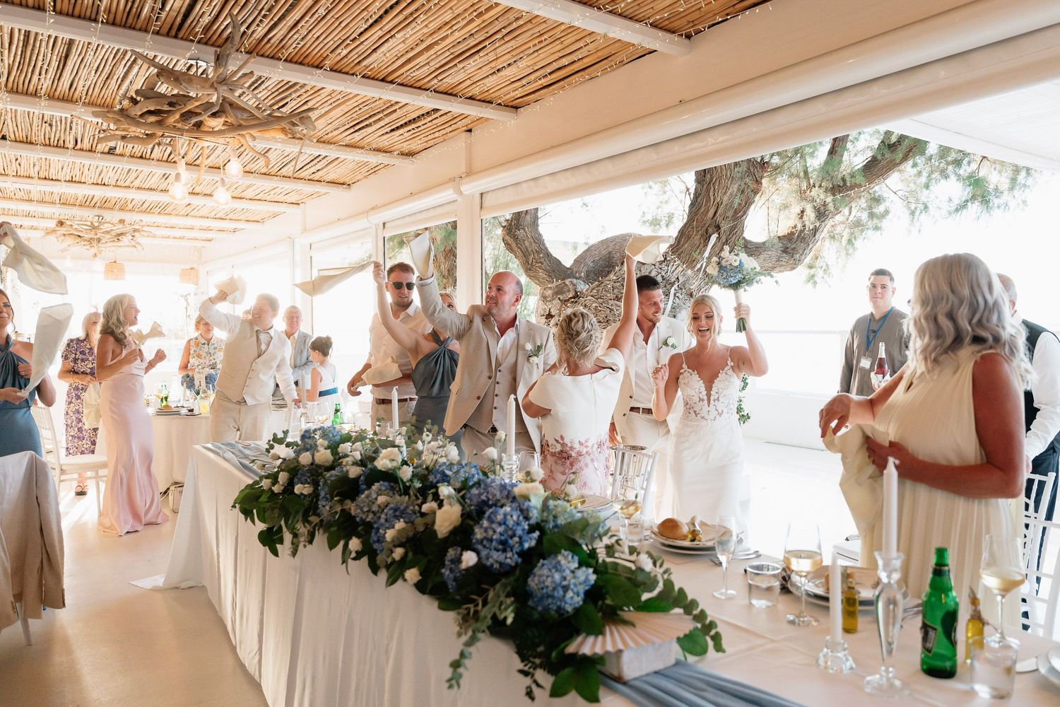 Wedding guests dance and celebrate in a rustic reception venue decorated with hanging lights and blue floral arrangements.