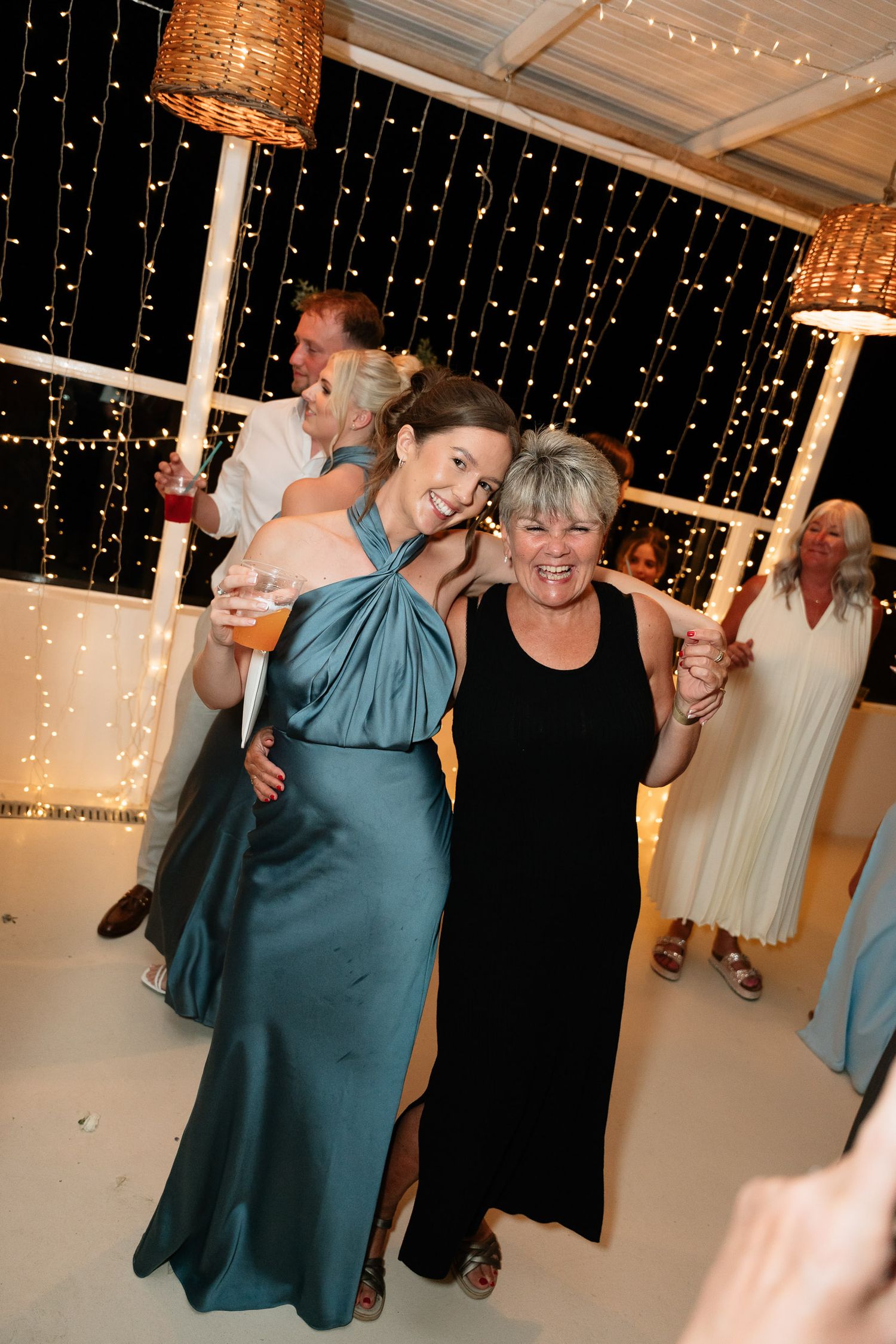 Wedding guests dancing and celebrating on a white dance floor under twinkling string lights and woven pendant lamps.
