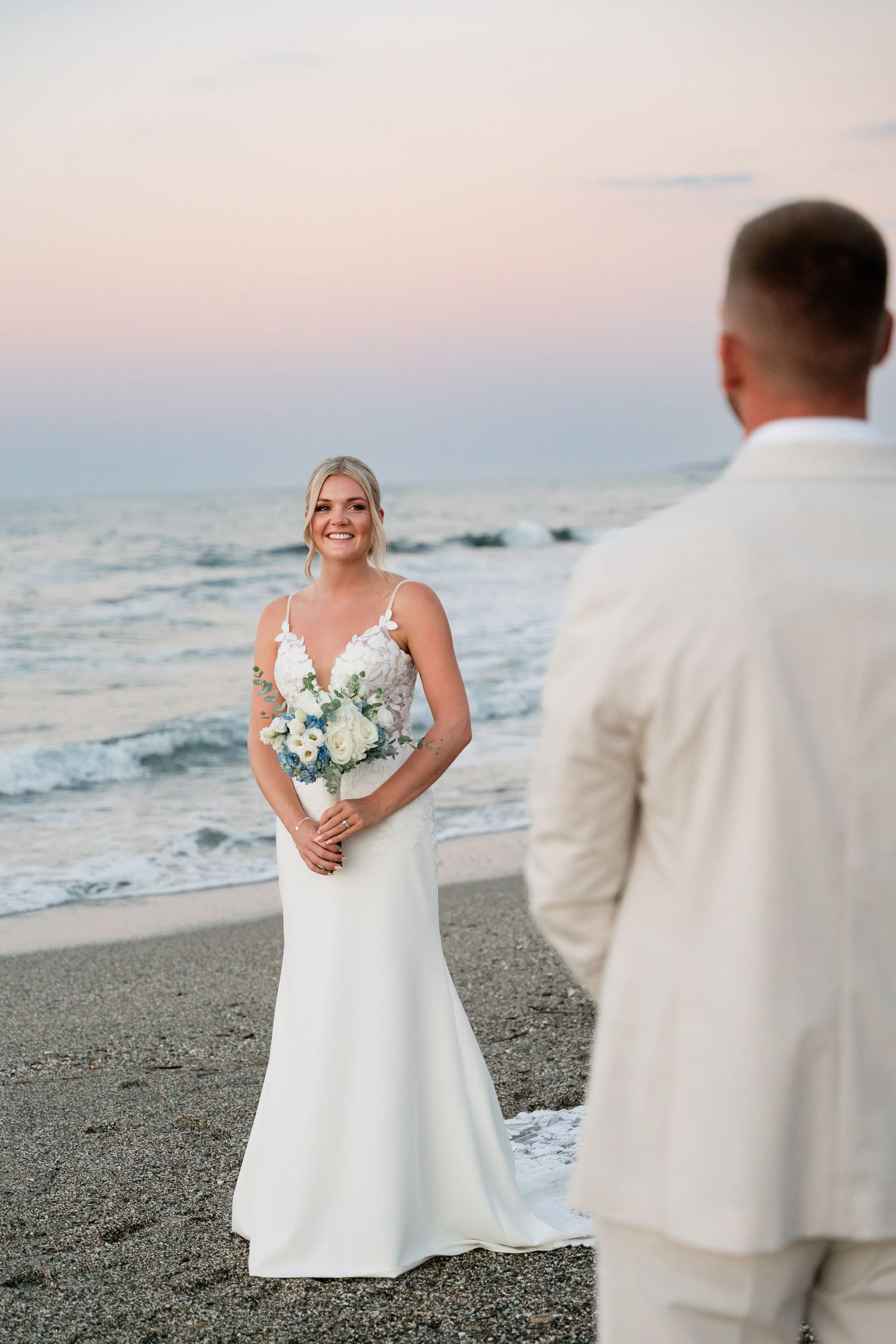A bride in a white wedding dress holds a bouquet while standing on a beach at sunset with waves in the background.