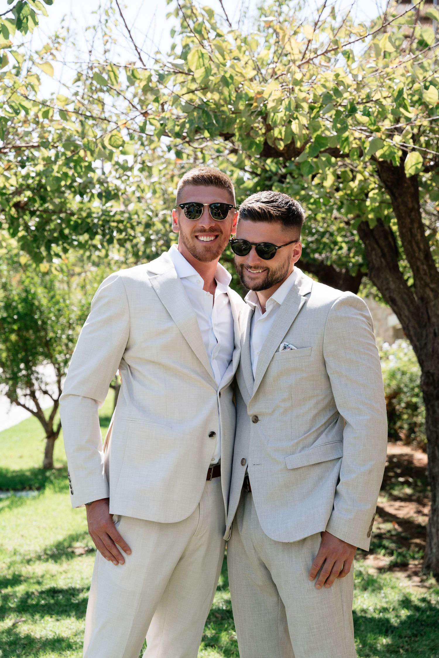 Two people in light beige suits and sunglasses pose together outdoors under flowering trees on a sunny day.
