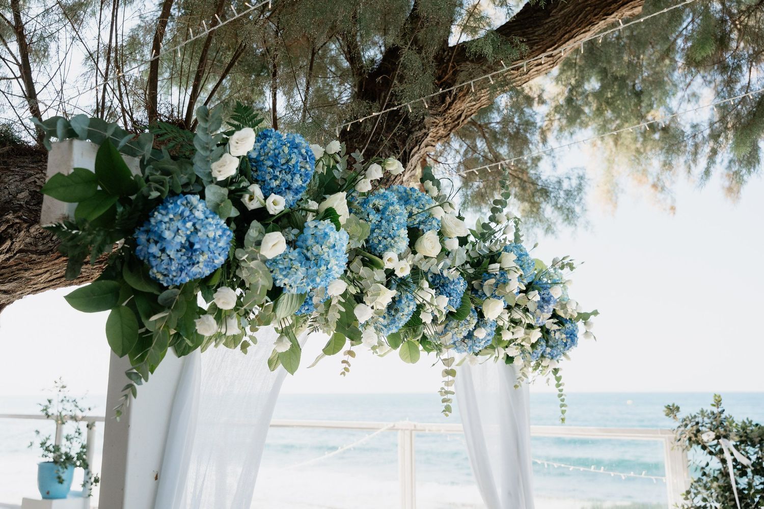 Decorative floral arch with blue hydrangeas and white flowers creates an elegant wedding ceremony backdrop on a beach.