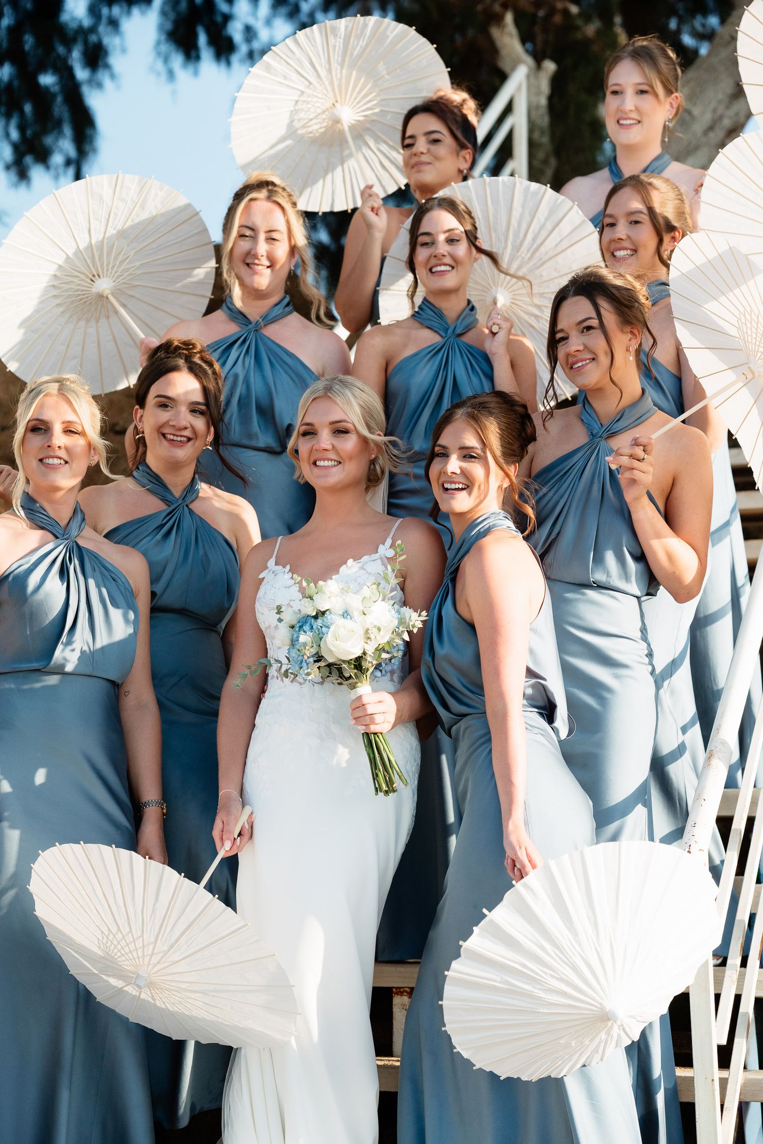 Wedding party poses on stairs with white paper parasols as bridesmaids wear matching blue dresses beside bride in white gown.