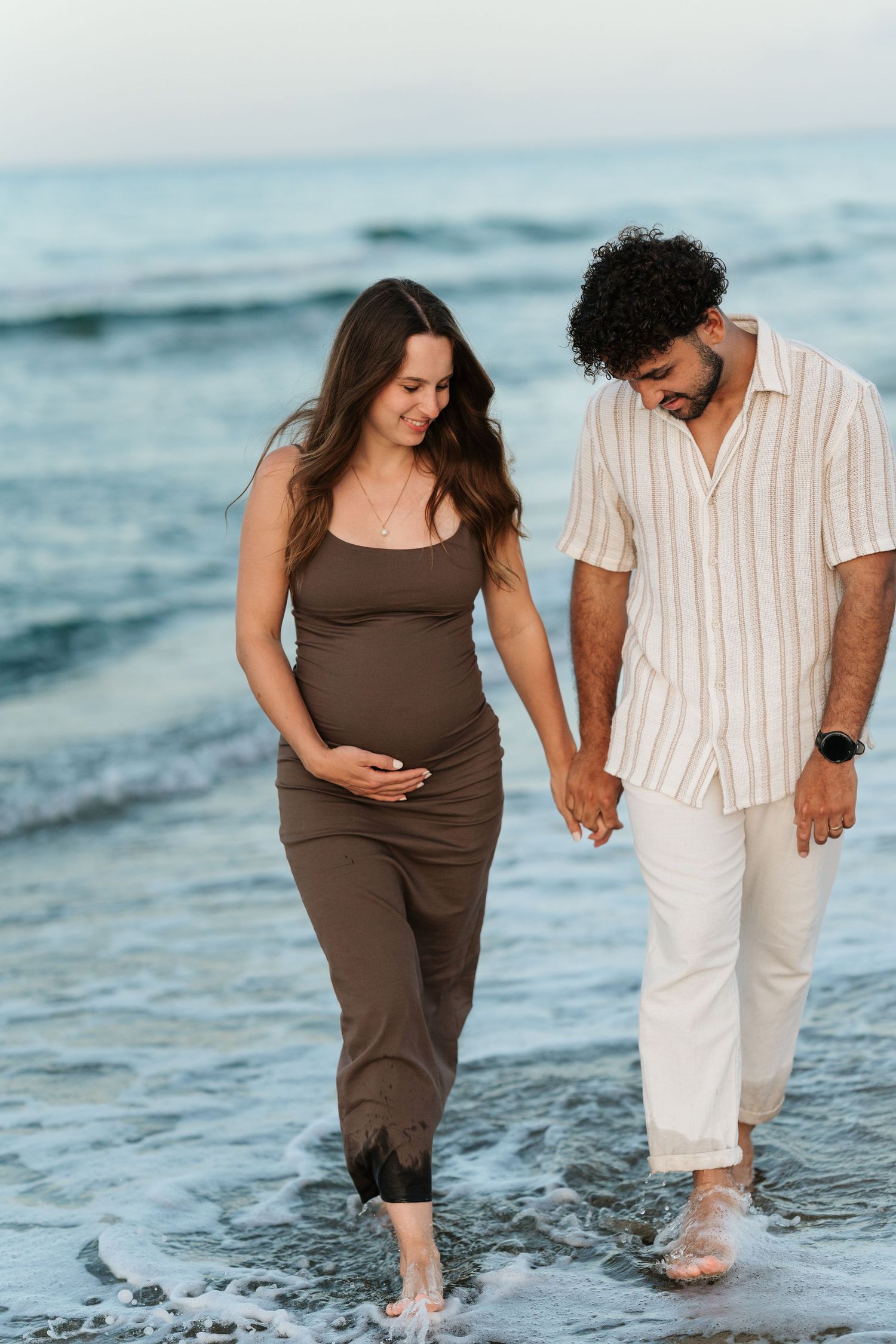 A couple walking together along the beach shoreline in matching brown and white outfits as waves lap at their feet.