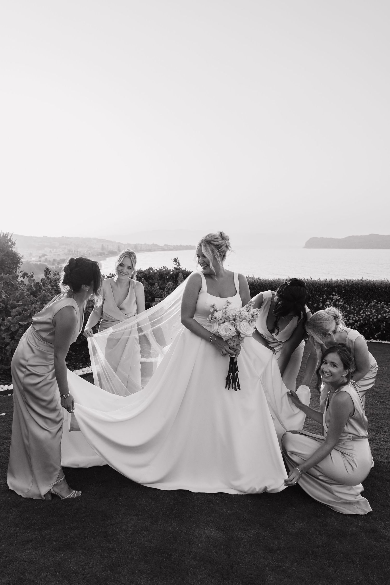 A wedding party in light colored dresses helps arrange the bride's flowing white gown on a scenic coastal lawn.
