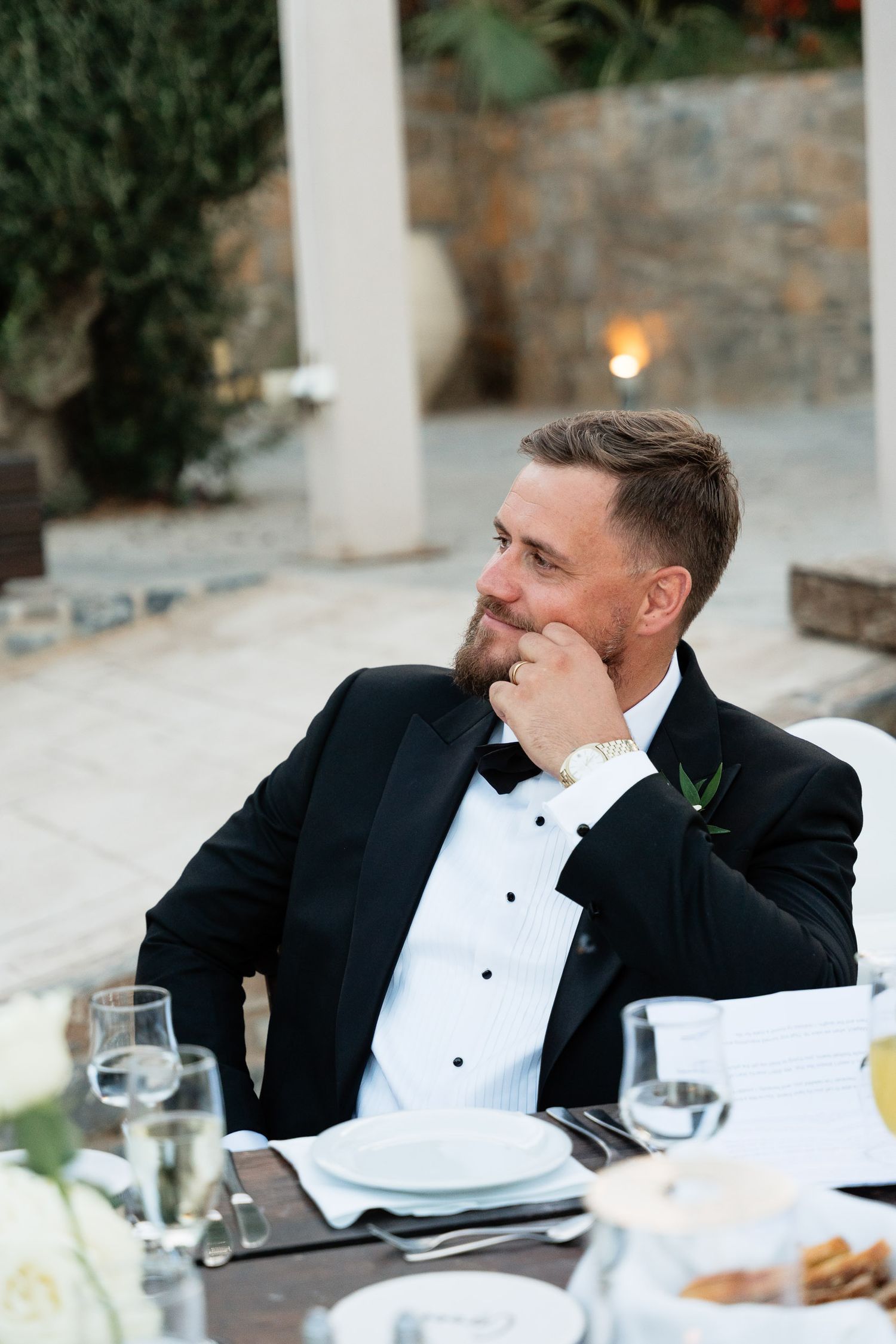 A person in a black tuxedo and white shirt sits at an elegant dining table with glassware and place settings.