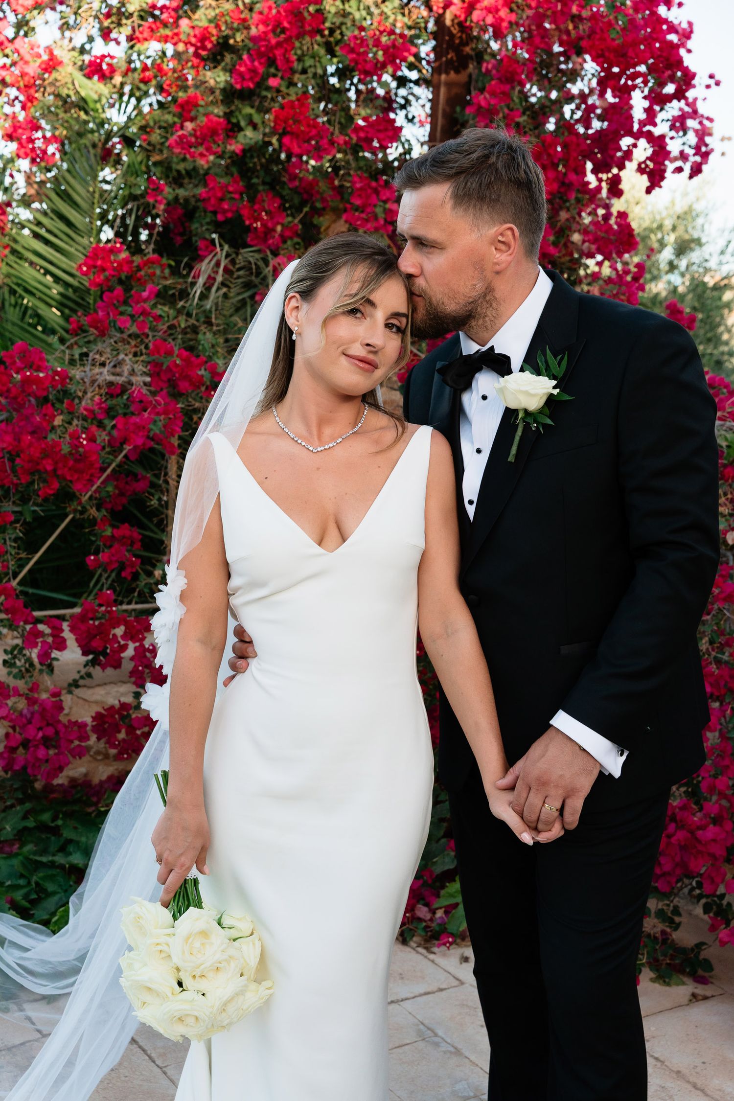 A romantic wedding couple poses together against vibrant pink bougainvillea flowers in an outdoor garden setting.