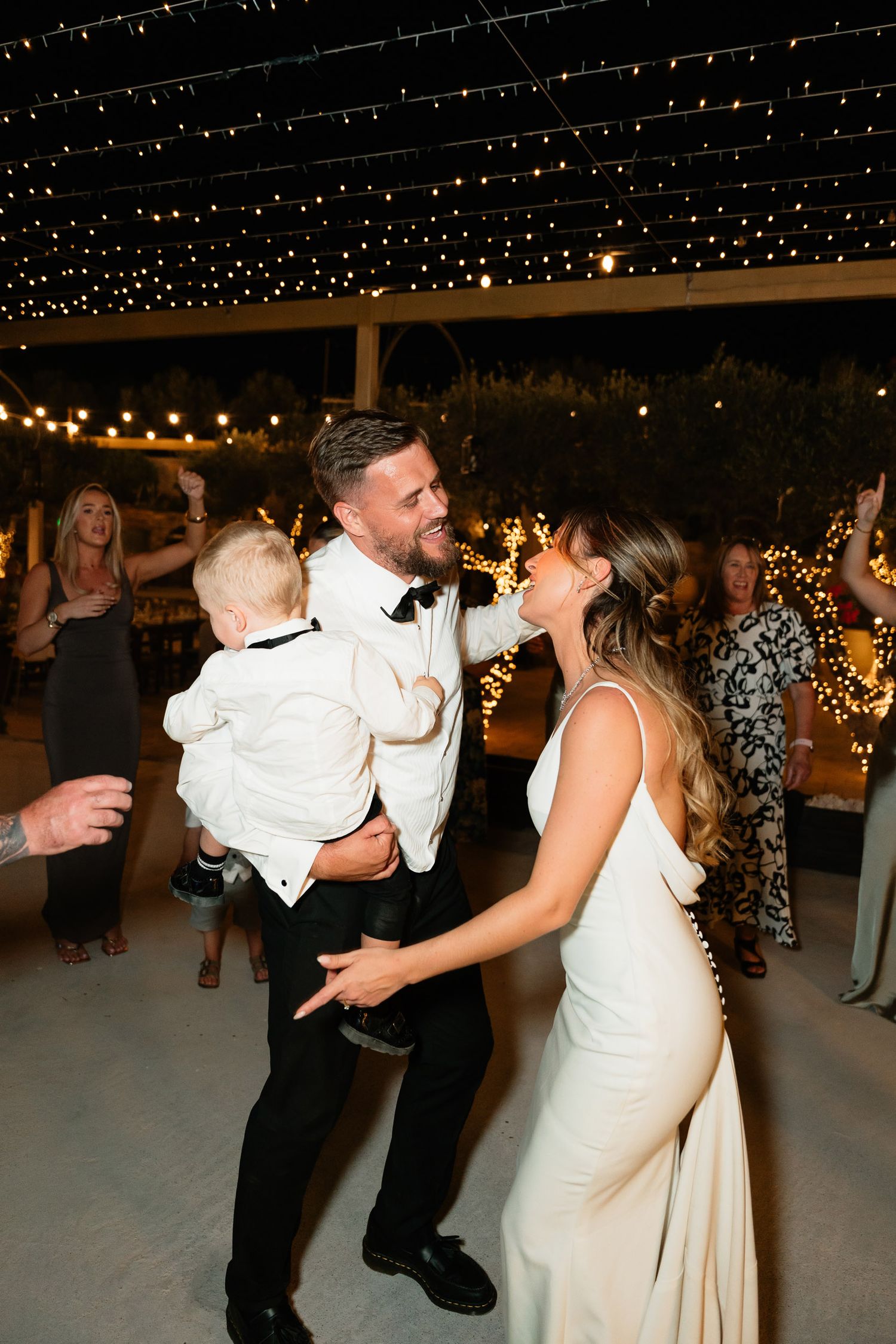 A couple dances together at a wedding reception under twinkling lights and a dark night sky.