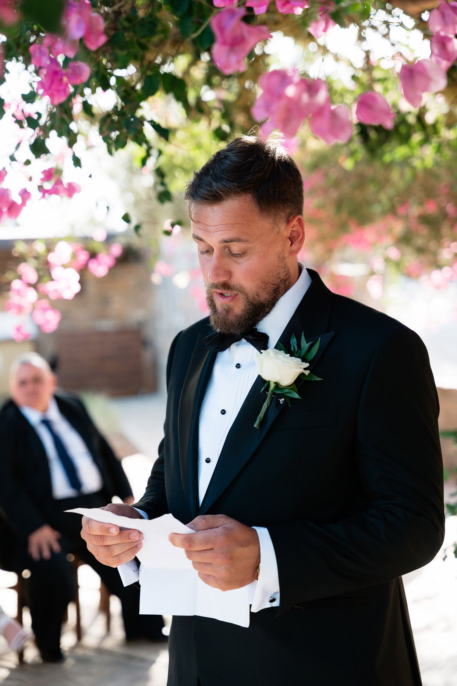 A groom in a black tuxedo reads a letter under pink flowering trees during an emotional wedding moment.