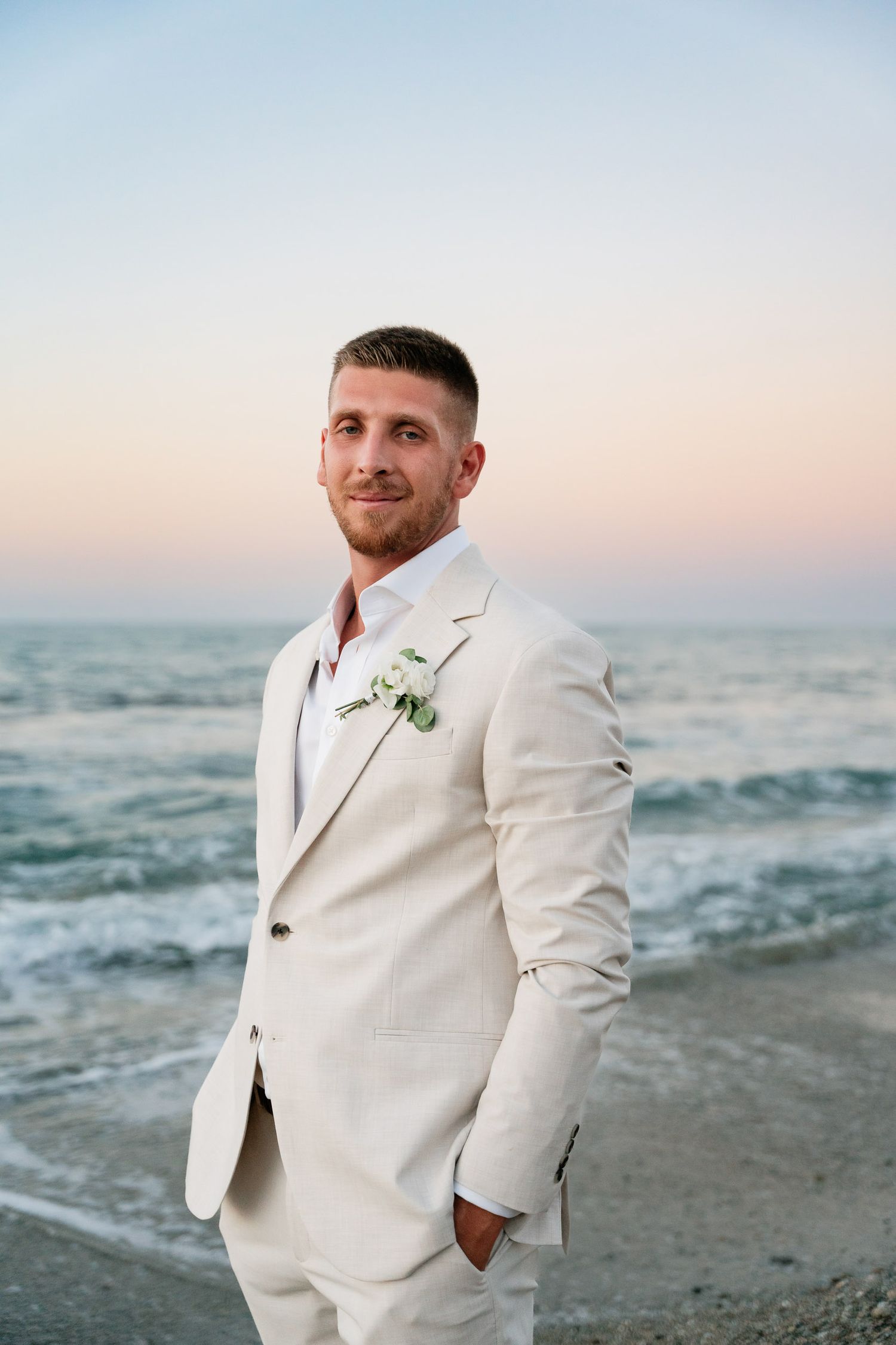 A groom in a cream-colored suit with boutonniere poses at sunset on a beach with ocean waves in the background.