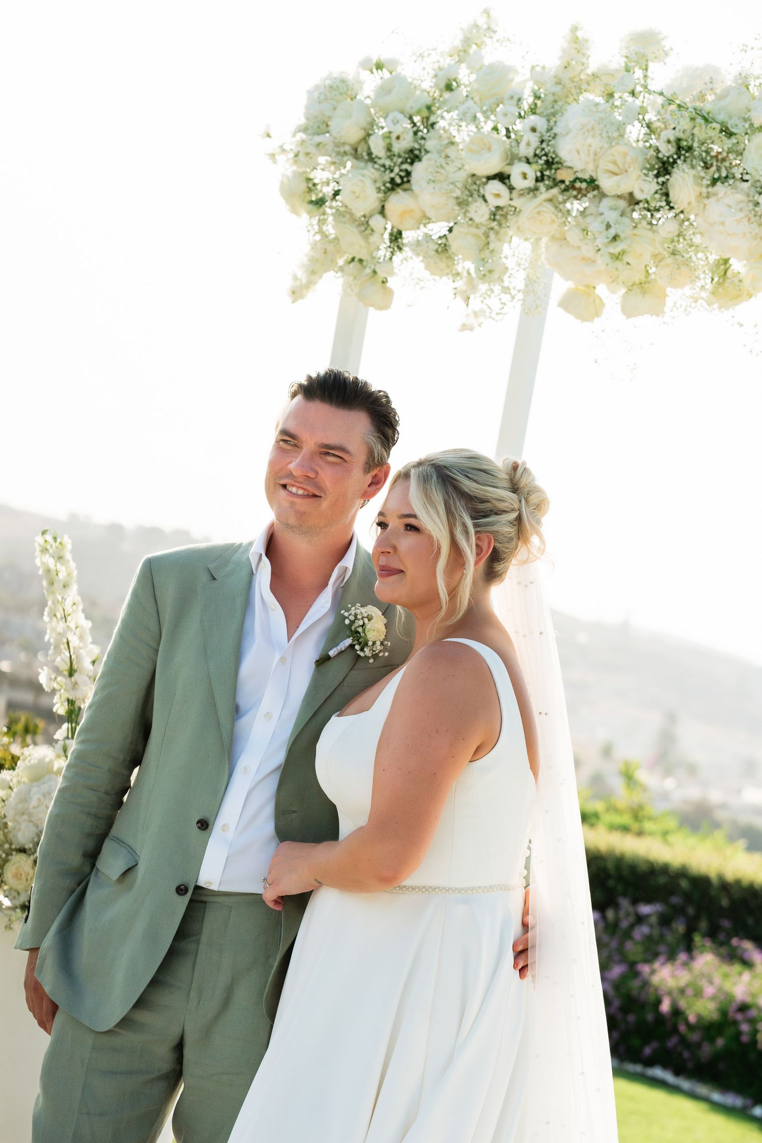 A couple shares joyful laughter during their outdoor wedding ceremony with white floral arrangements in the background.