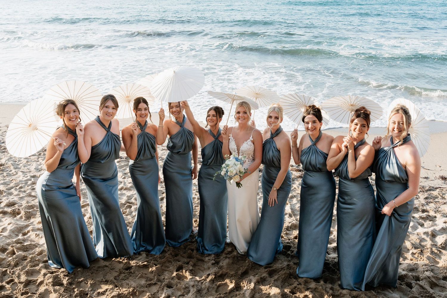 A group of bridesmaids in matching blue dresses pose together on a beach with ocean waves in the background.