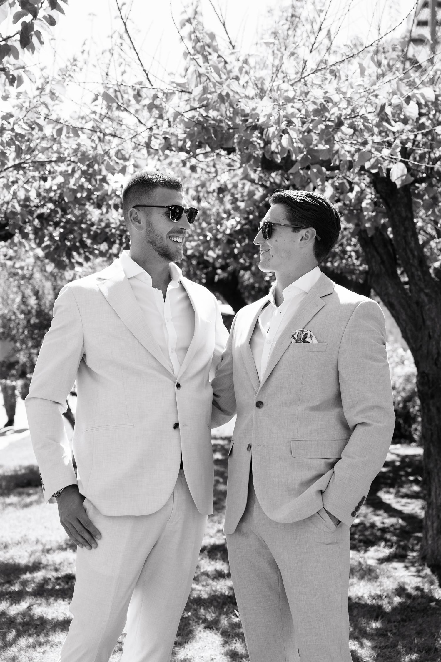 A group of groomsmen in light colored suits pose casually together outdoors under blooming trees in a candid moment.
