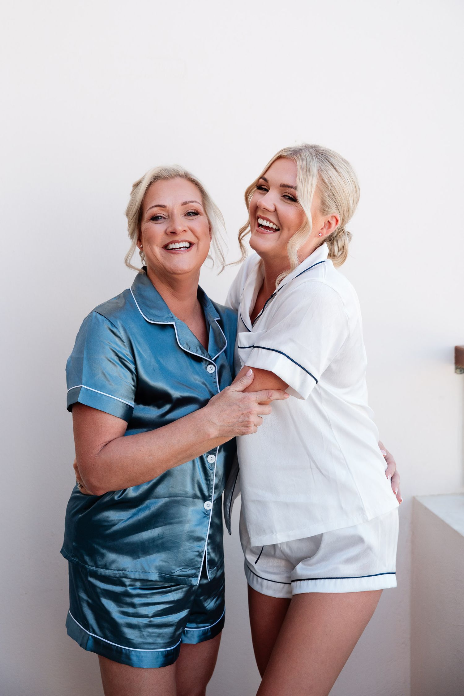 Two women wearing satin pajama sets laugh together against a white wall.