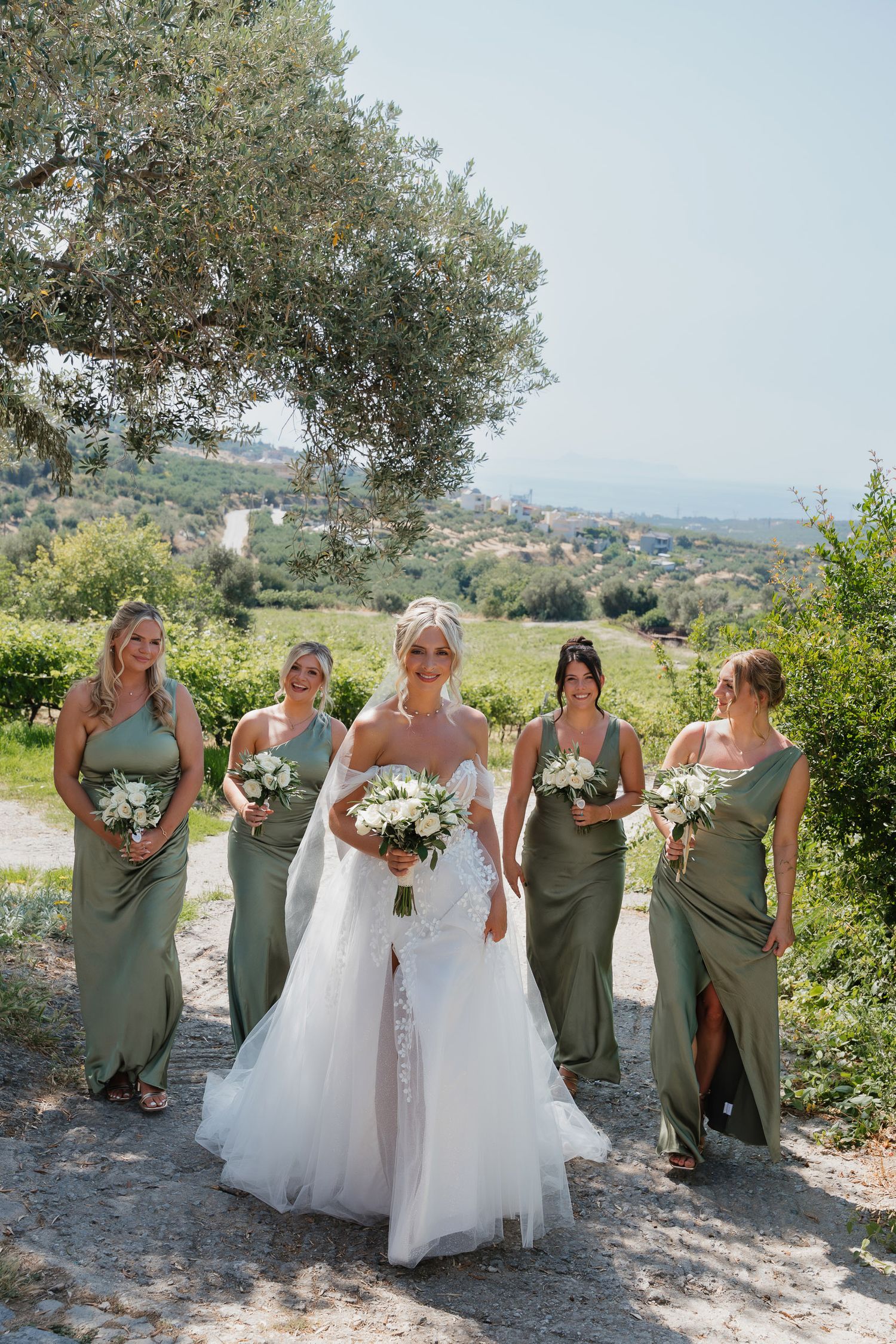 A bridal party in sage green dresses and white gowns poses together on a sunny hillside with scenic views.