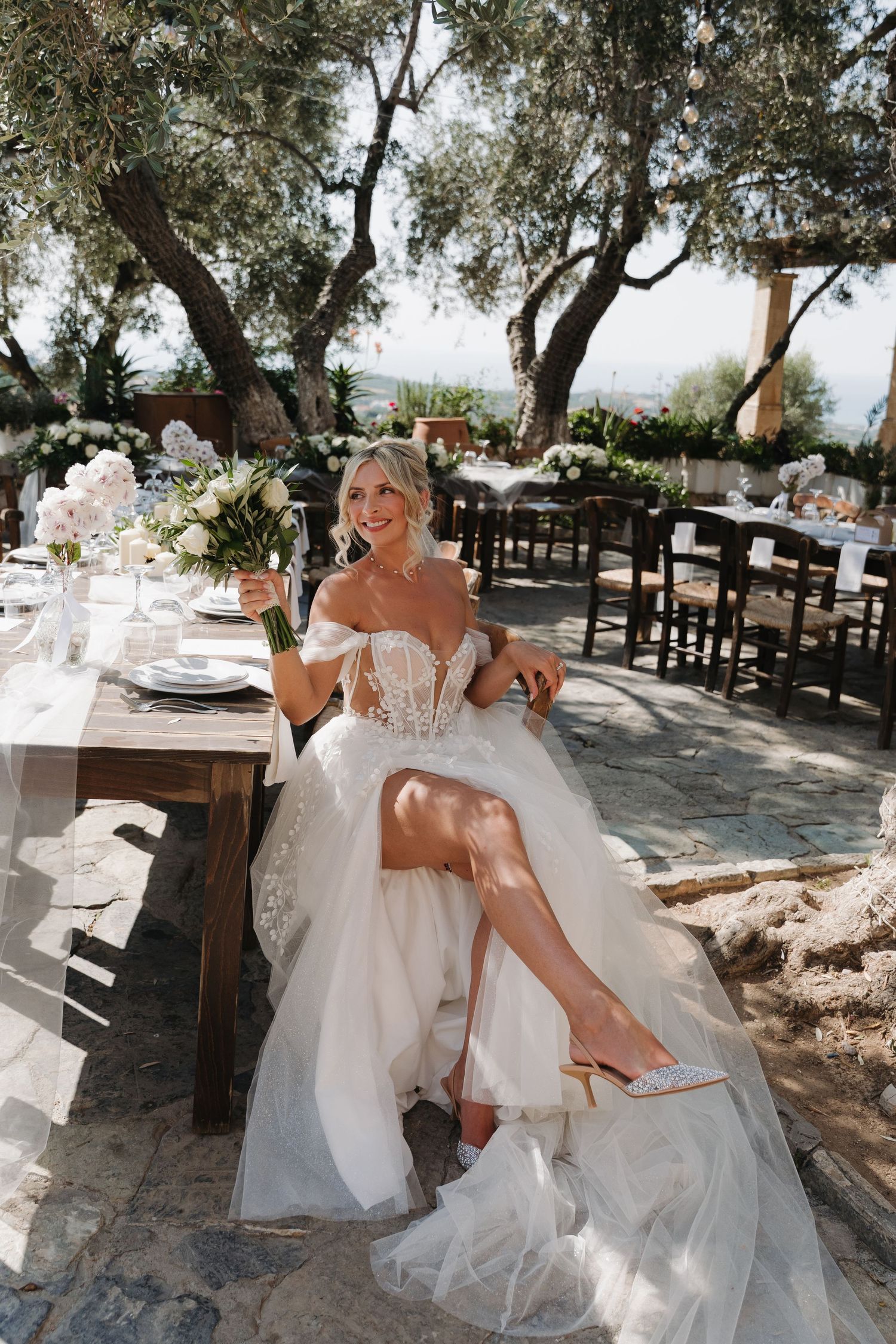 A bride in a white off-shoulder wedding gown poses outdoors under olive trees at a rustic venue setting.