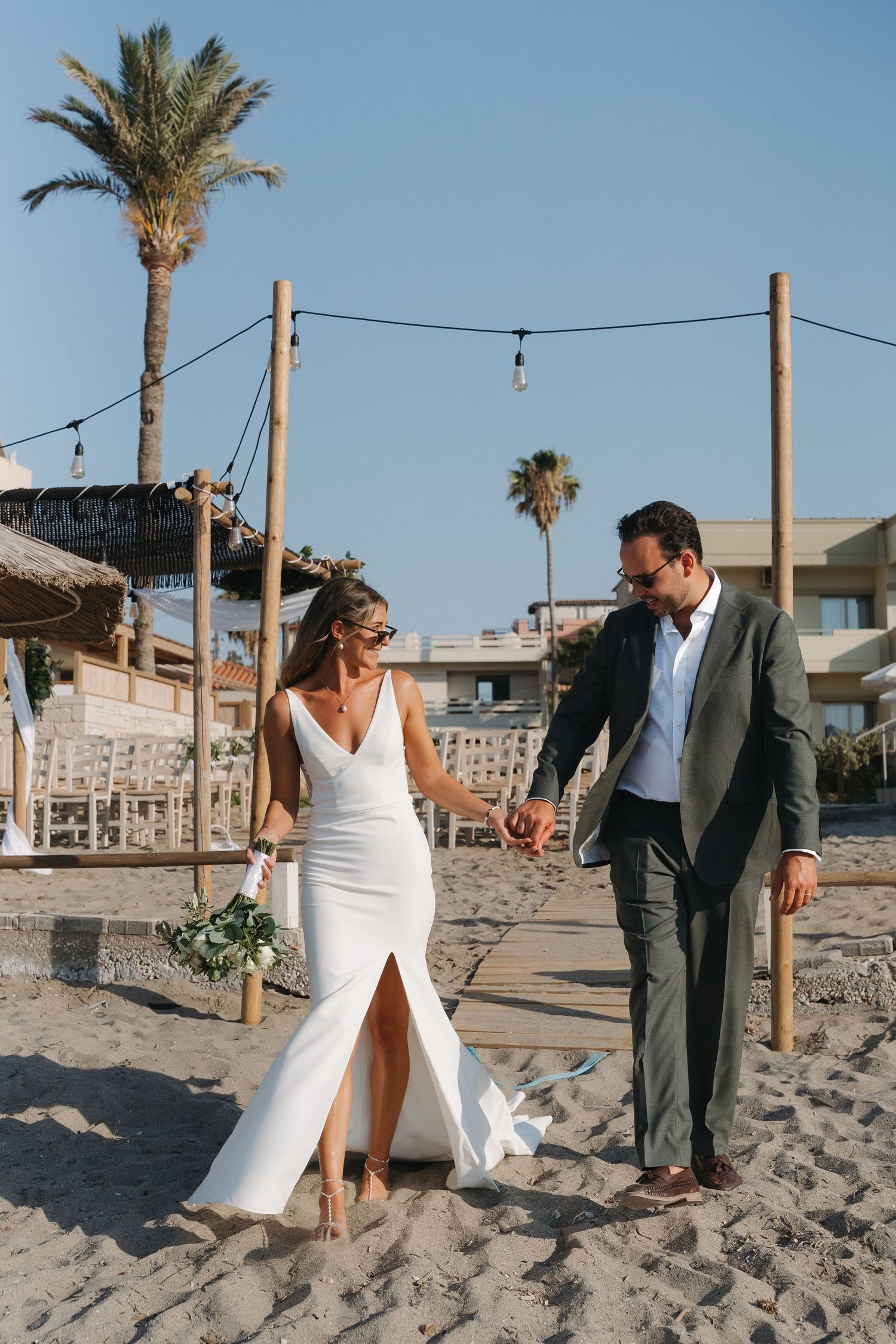 Newlyweds walk hand in hand on sandy beach with palm trees and string lights overhead during sunset wedding.