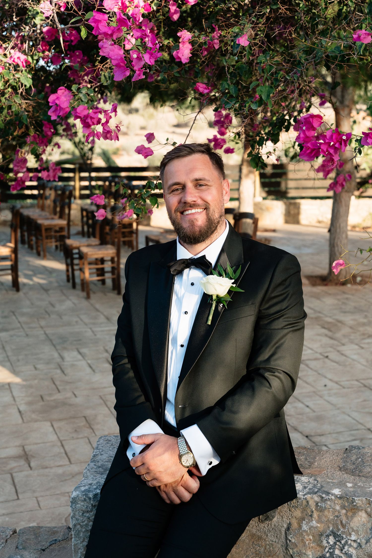 A groom in a black tuxedo sits outdoors under vibrant pink bougainvillea flowers on a stone patio.