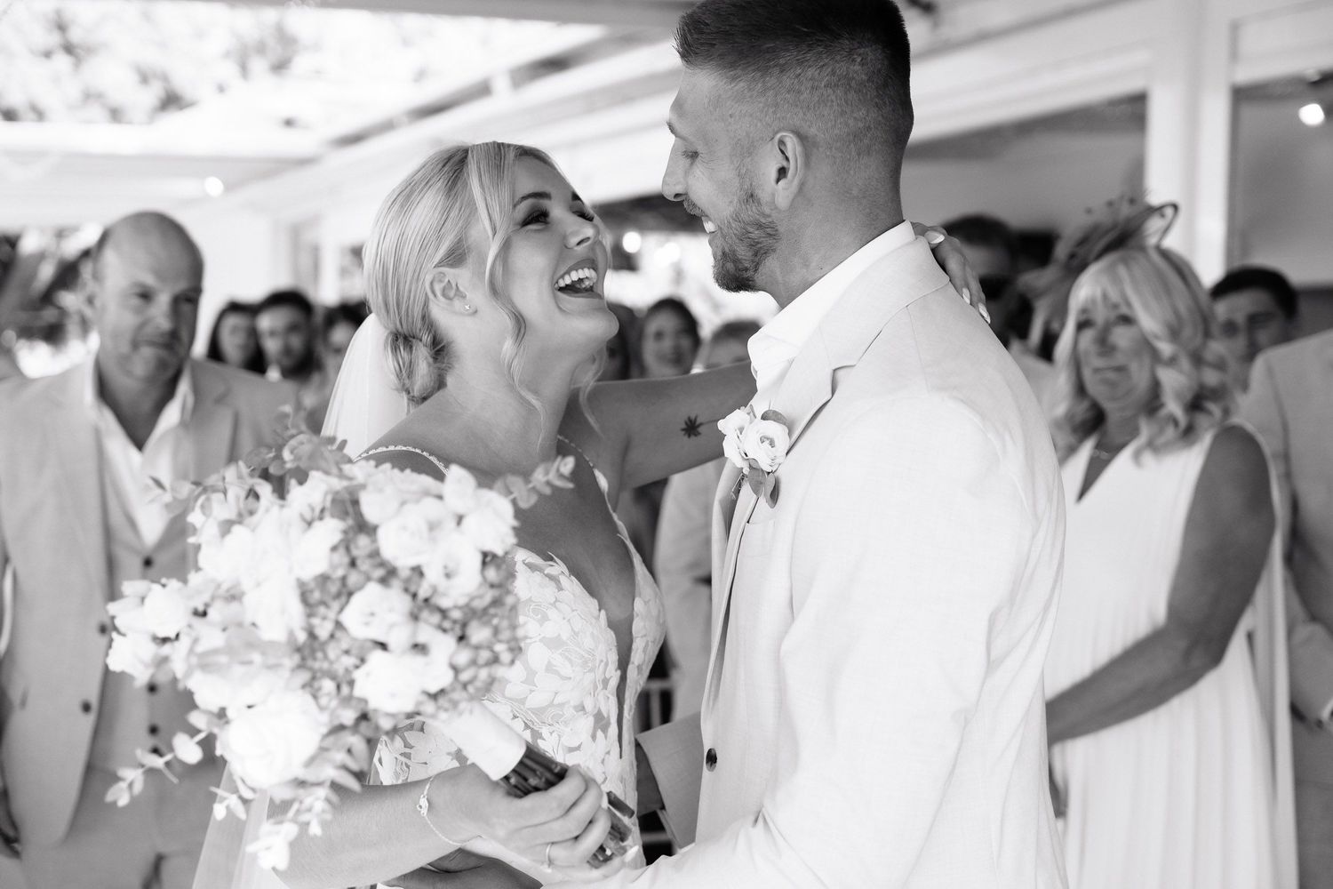 A joyful black and white wedding moment with the newlyweds laughing together as the bride holds a white floral bouquet.