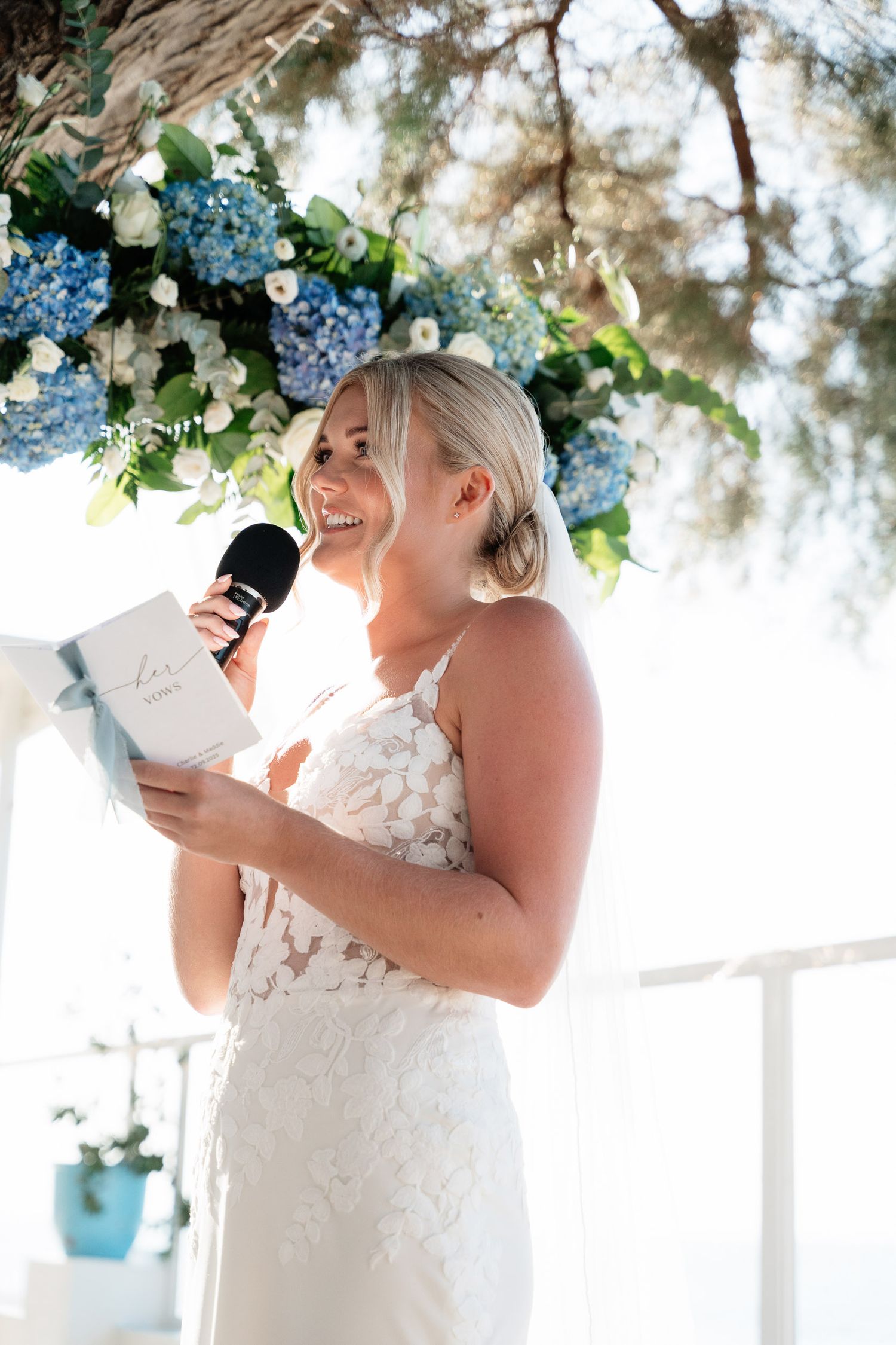 A bride in a white lace dress laughs joyfully while holding a drink during an outdoor wedding reception.