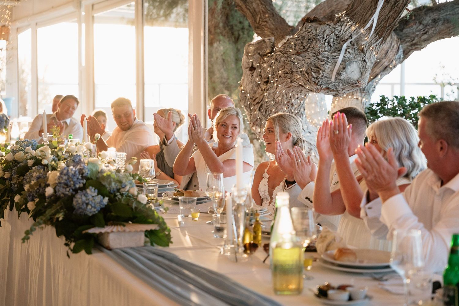 Wedding guests in white attire raise their glasses in celebration during an elegant outdoor reception dinner.