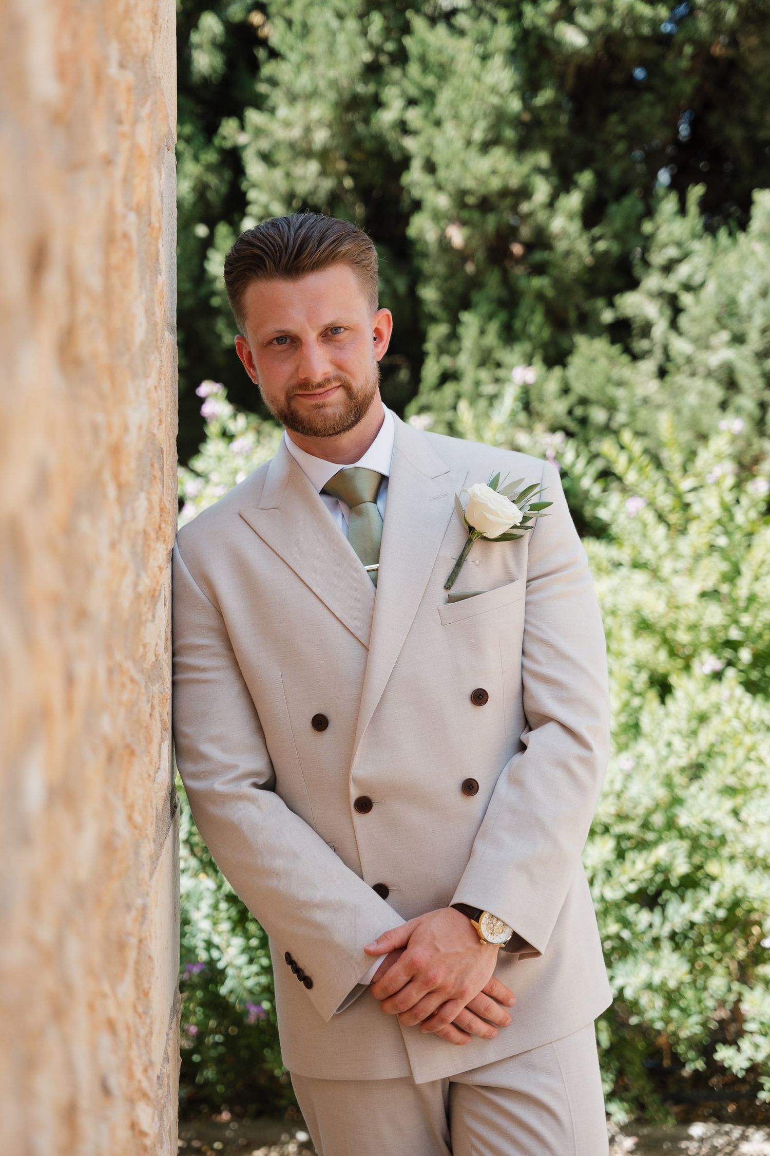 A person in a light beige double-breasted suit stands outdoors against a green foliage background.