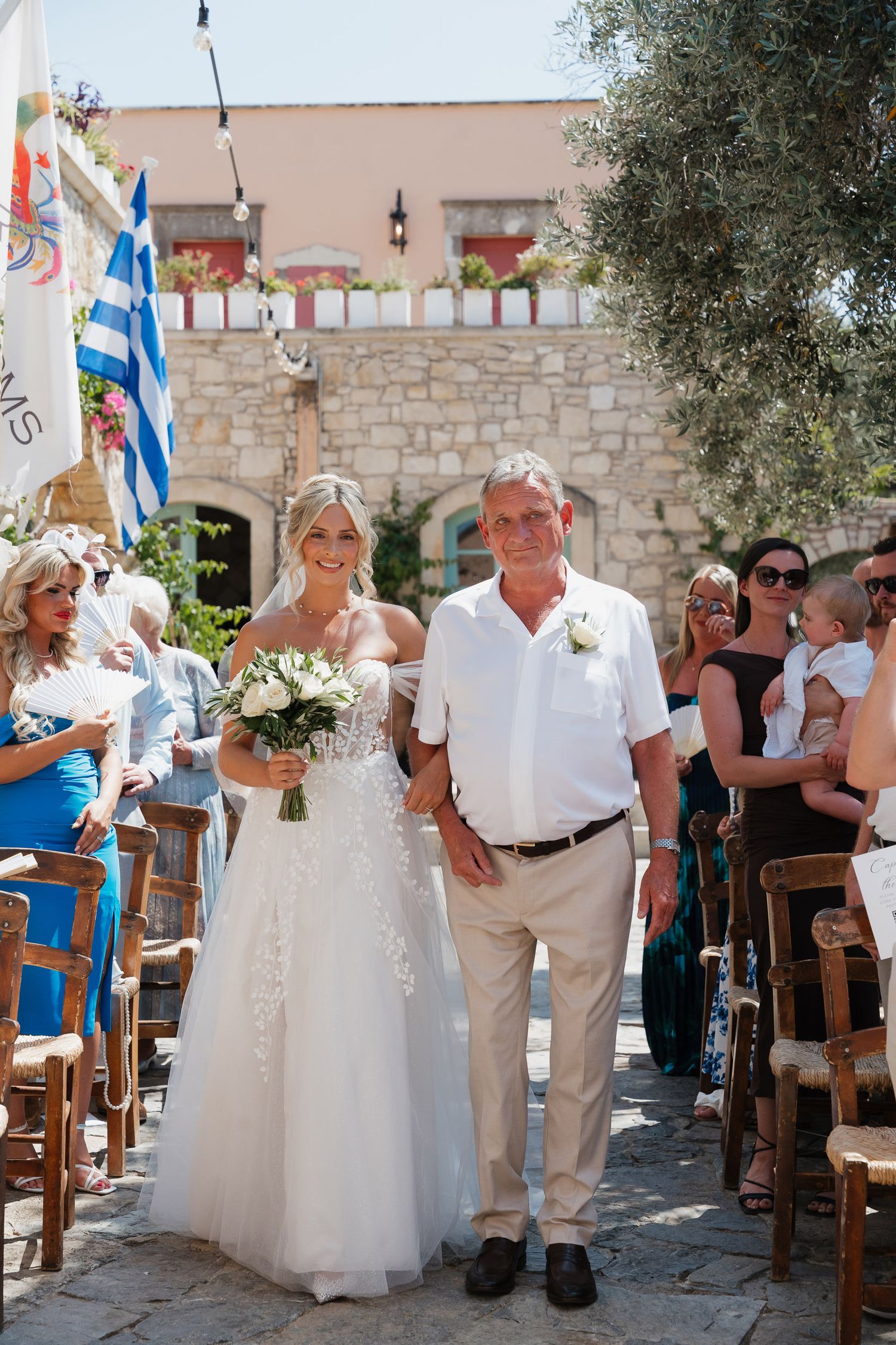 Wedding party guests in white attire dance and celebrate at a rustic Mediterranean stone venue with olive trees.