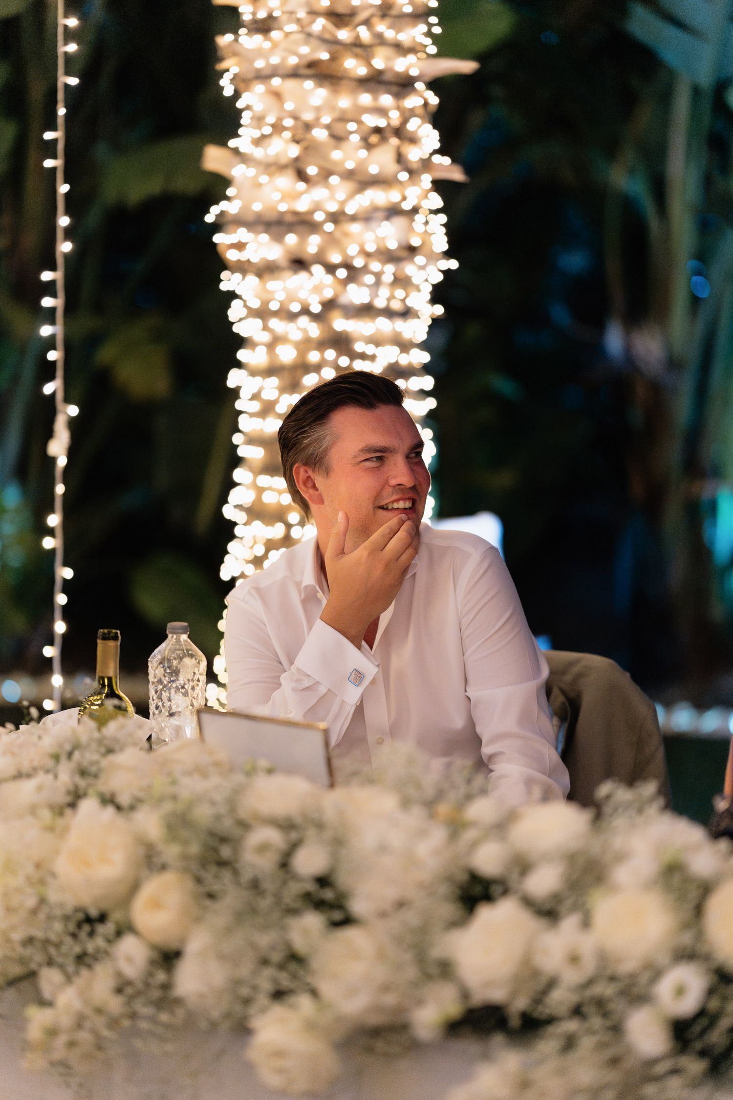 A person laughs while sitting at an elegant table decorated with white flowers and twinkling lights above.