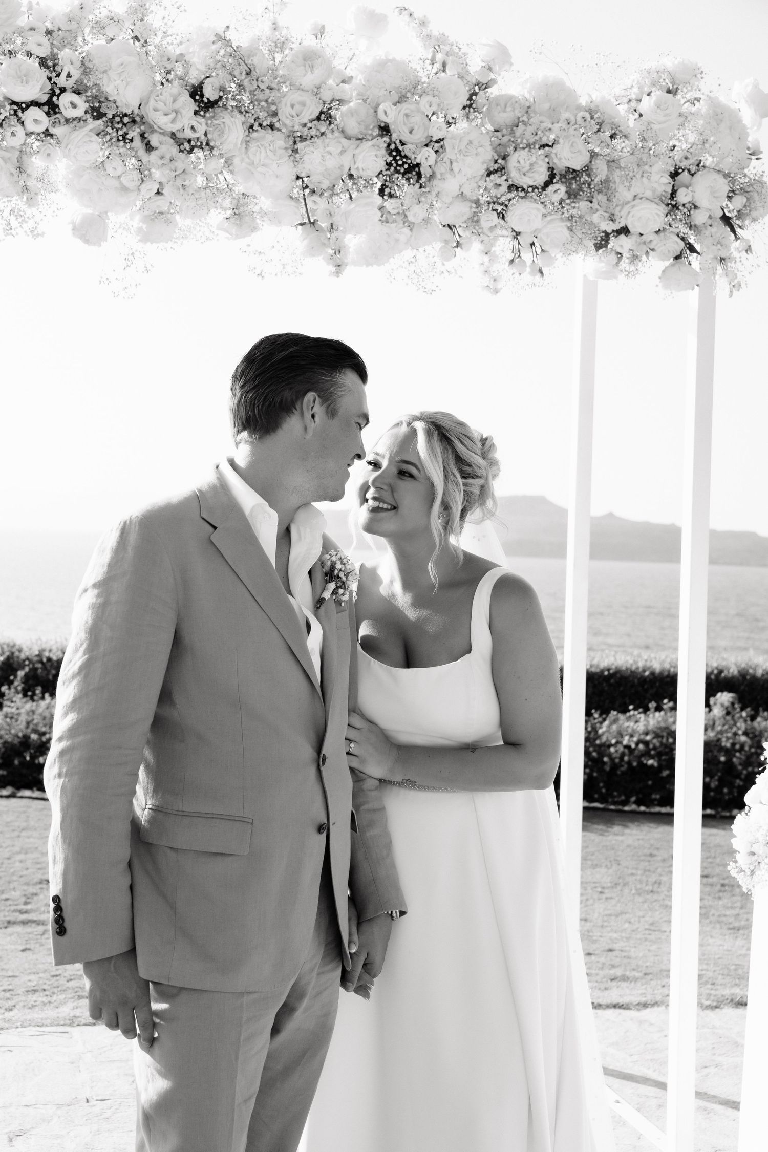 A romantic black and white wedding photo sequence showing tender moments between a couple by white pillars near the ocean.