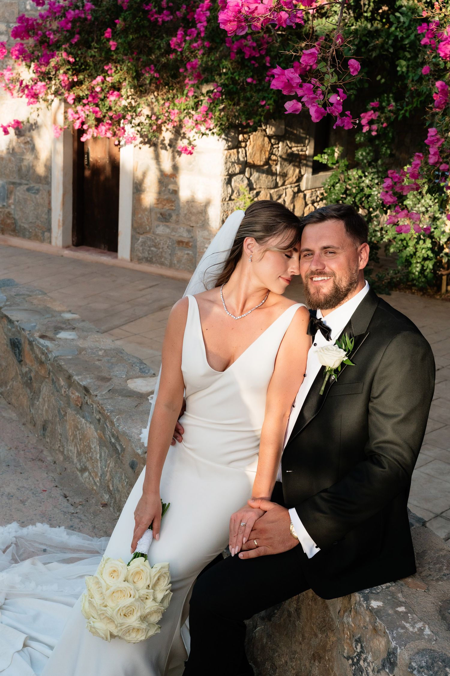 A romantic wedding couple shares intimate moments under vibrant pink bougainvillea flowers on stone steps.
