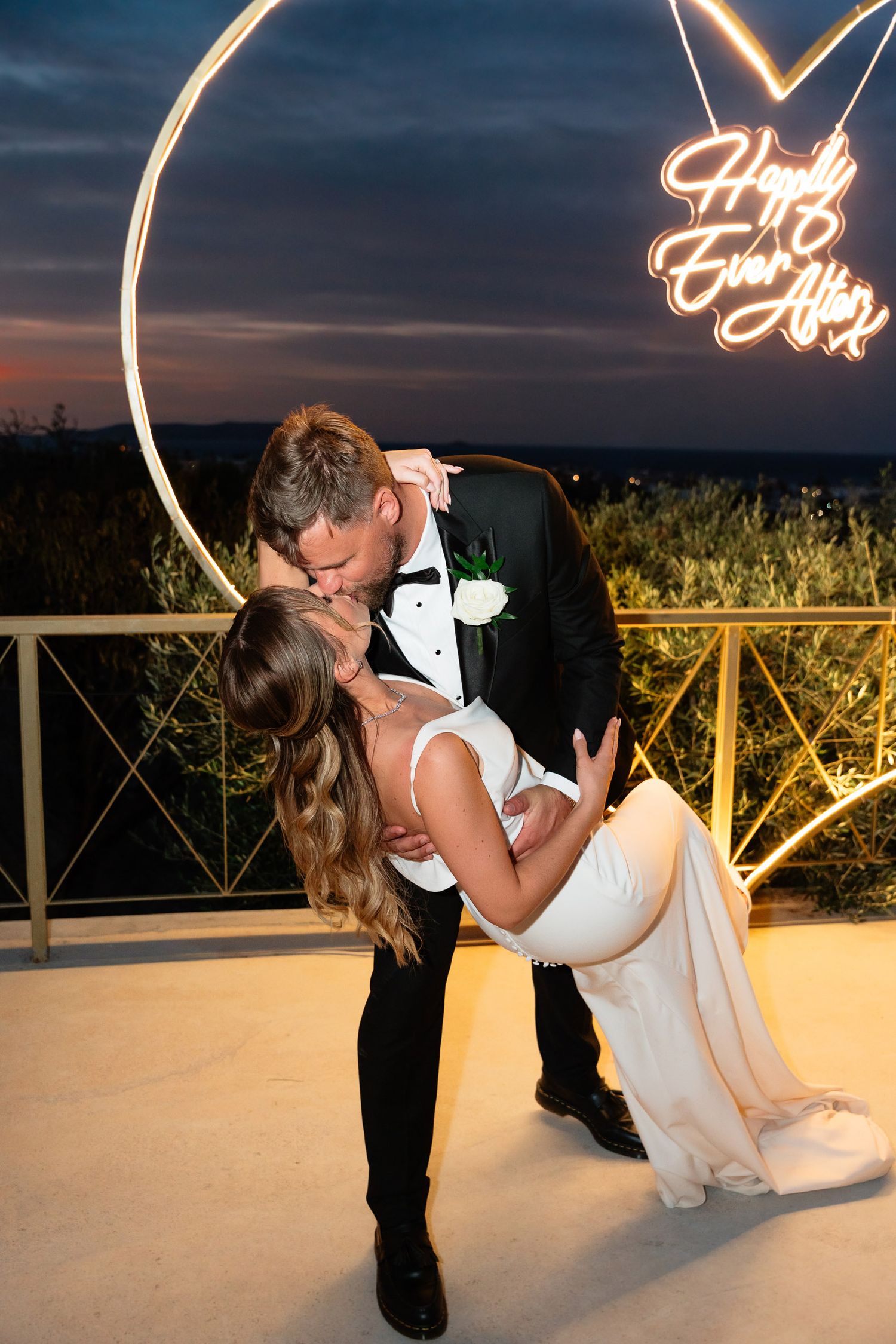 A romantic wedding couple shares a dramatic dip kiss on an illuminated dance floor at dusk.
