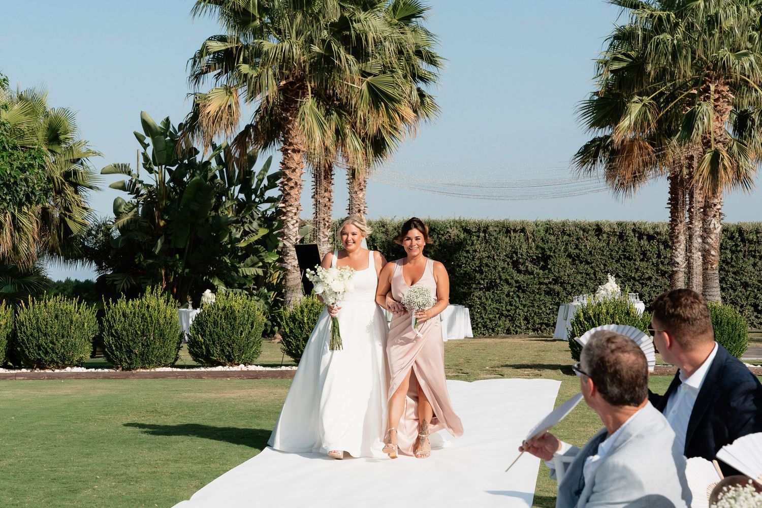 A scenic outdoor wedding ceremony takes place on a grassy lawn surrounded by palm trees on a sunny day.