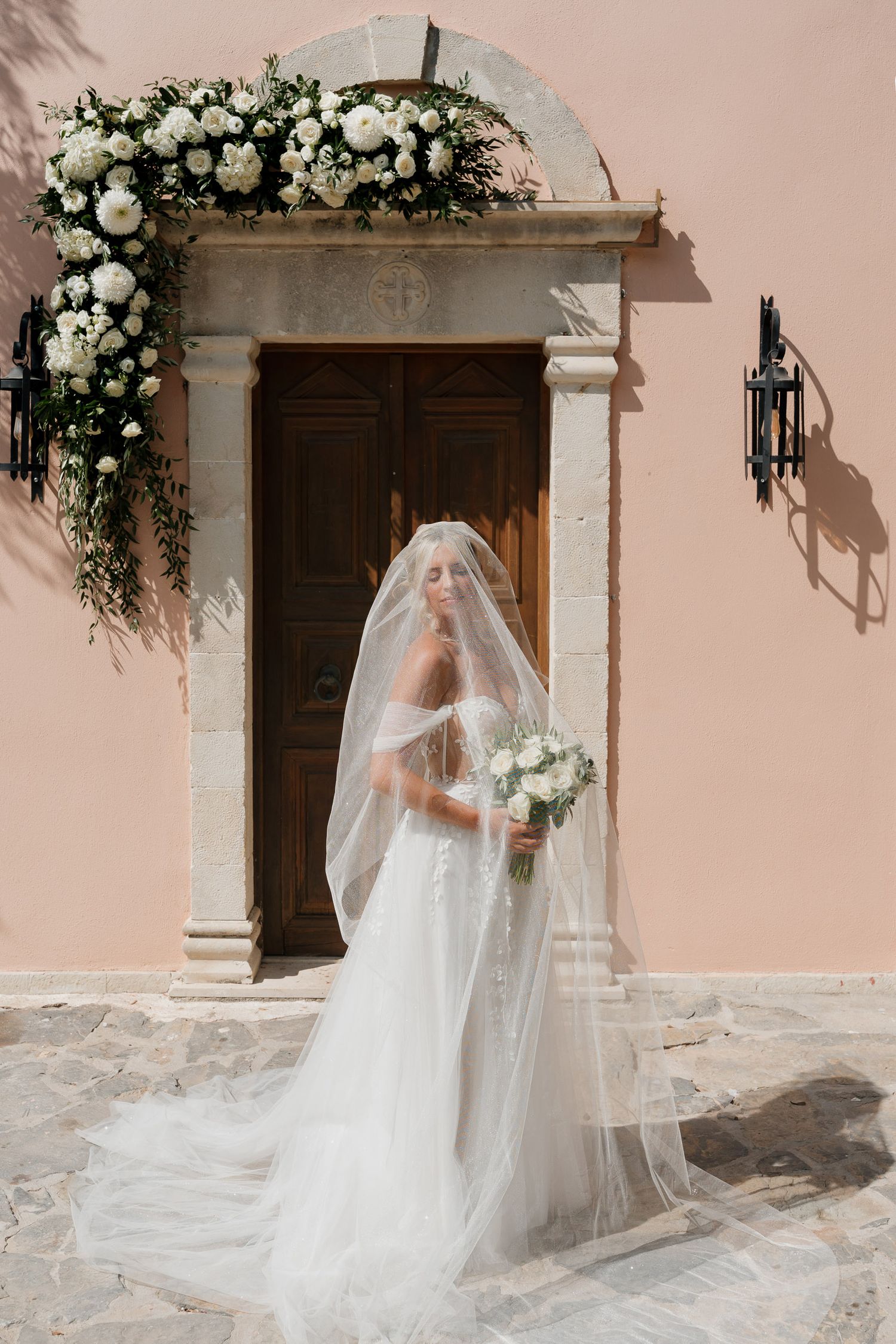 Elegant bridal portraits in front of a blush pink wall with white floral arrangements and wooden doors.