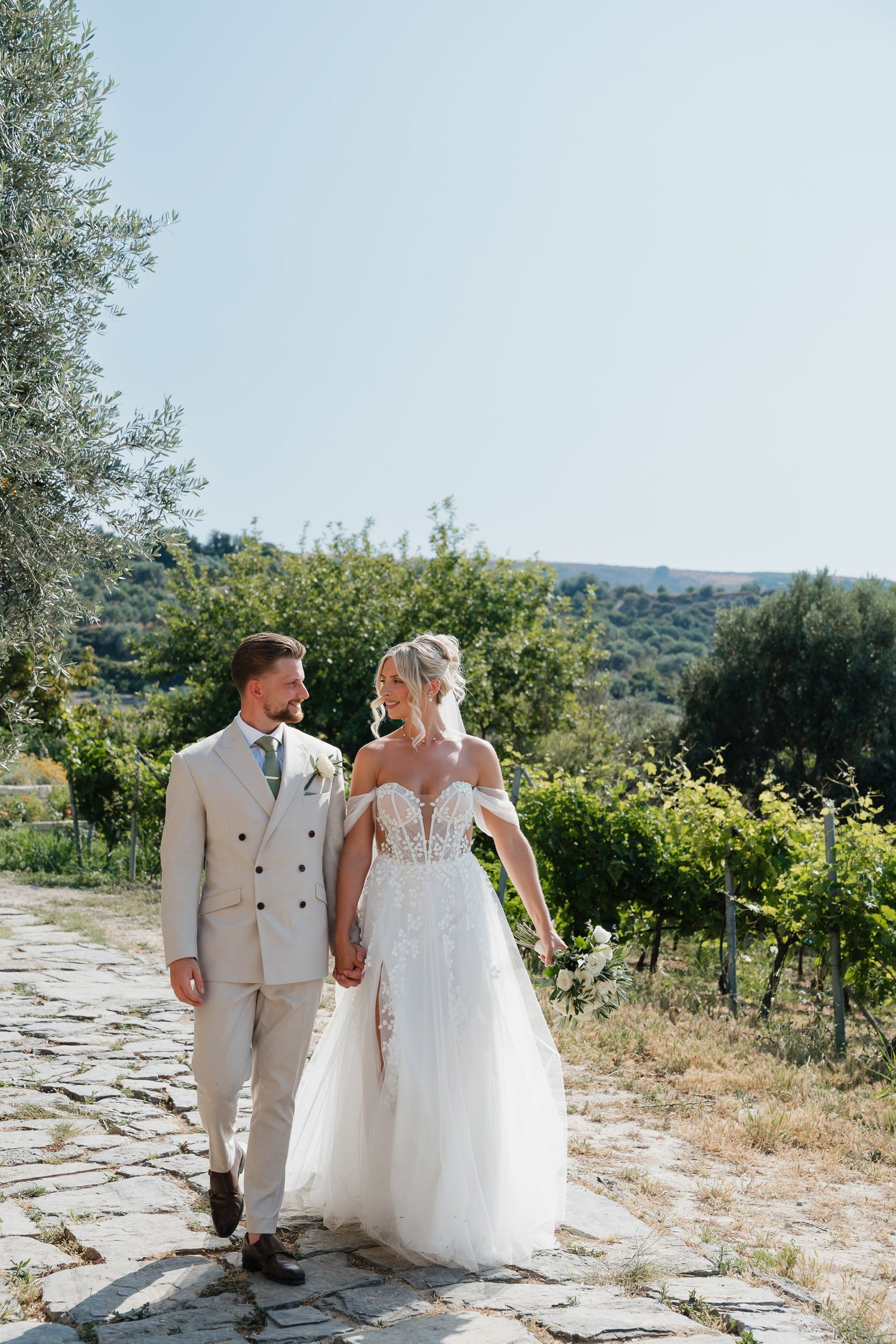 A couple in wedding attire walks together on a stone path surrounded by scenic countryside views.