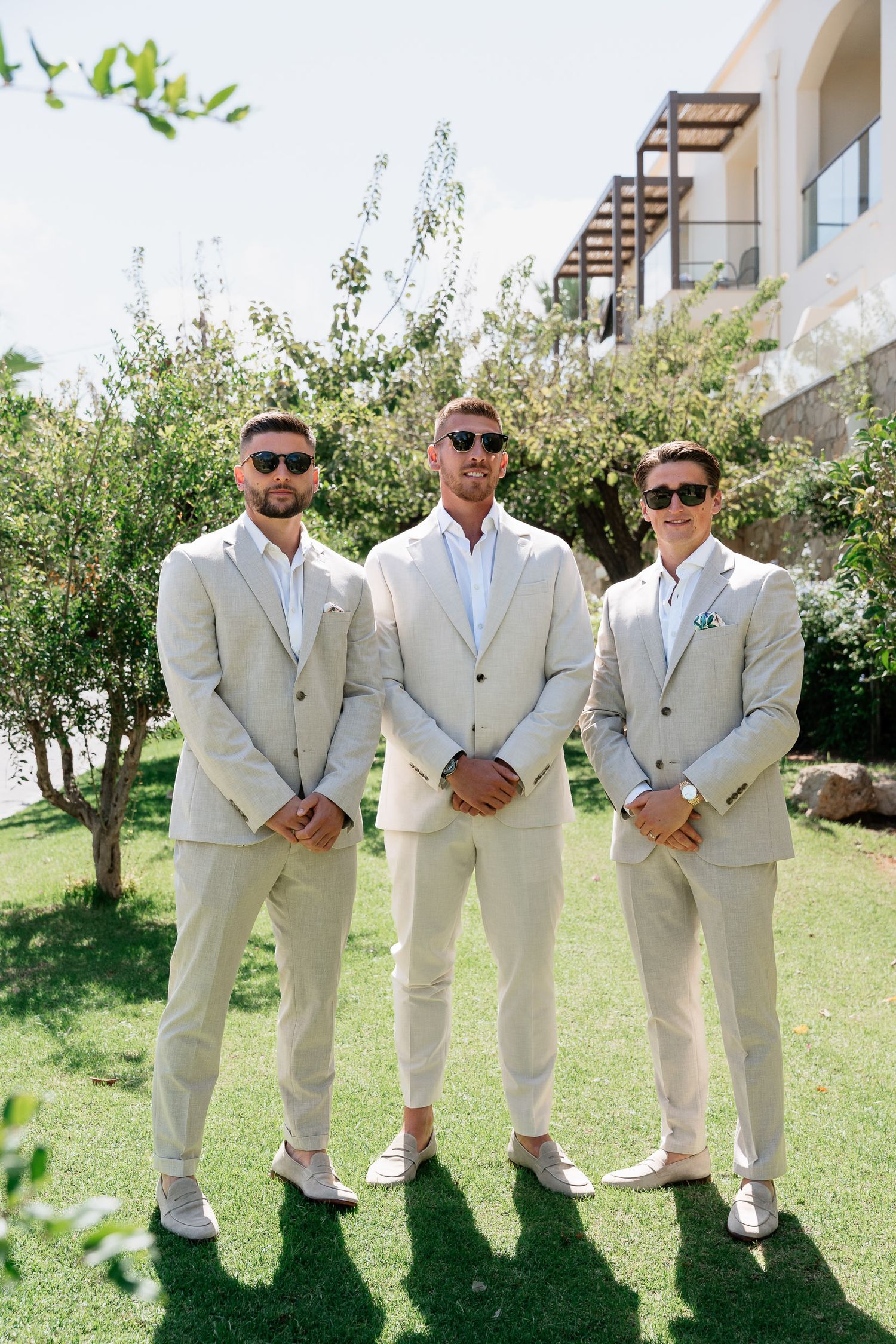 Groomsmen in matching light beige suits and sunglasses pose together on a sunny lawn with greenery in the background.