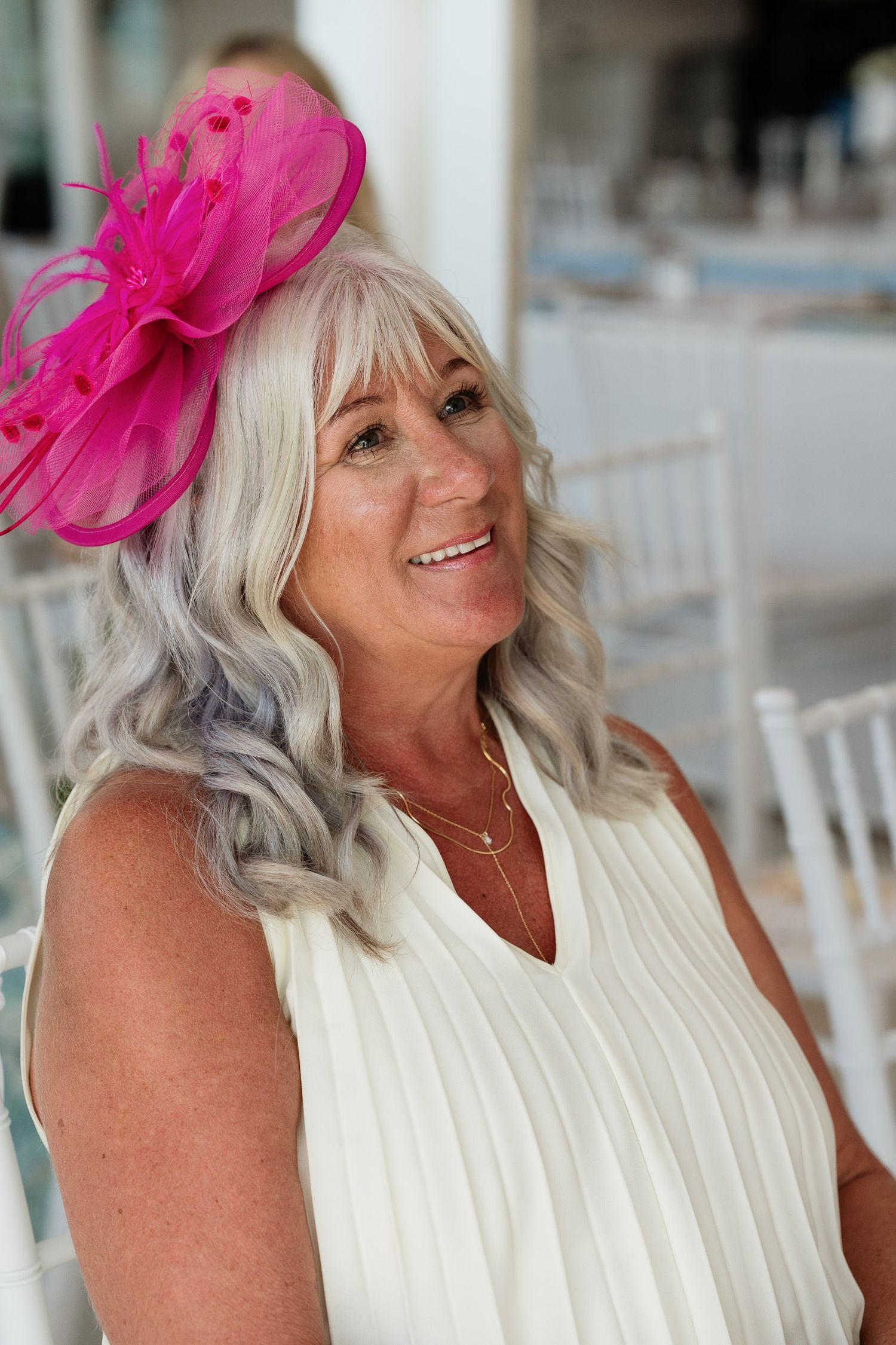 A person wearing a white sleeveless top and bright pink fascinator smiles while sitting at a table.