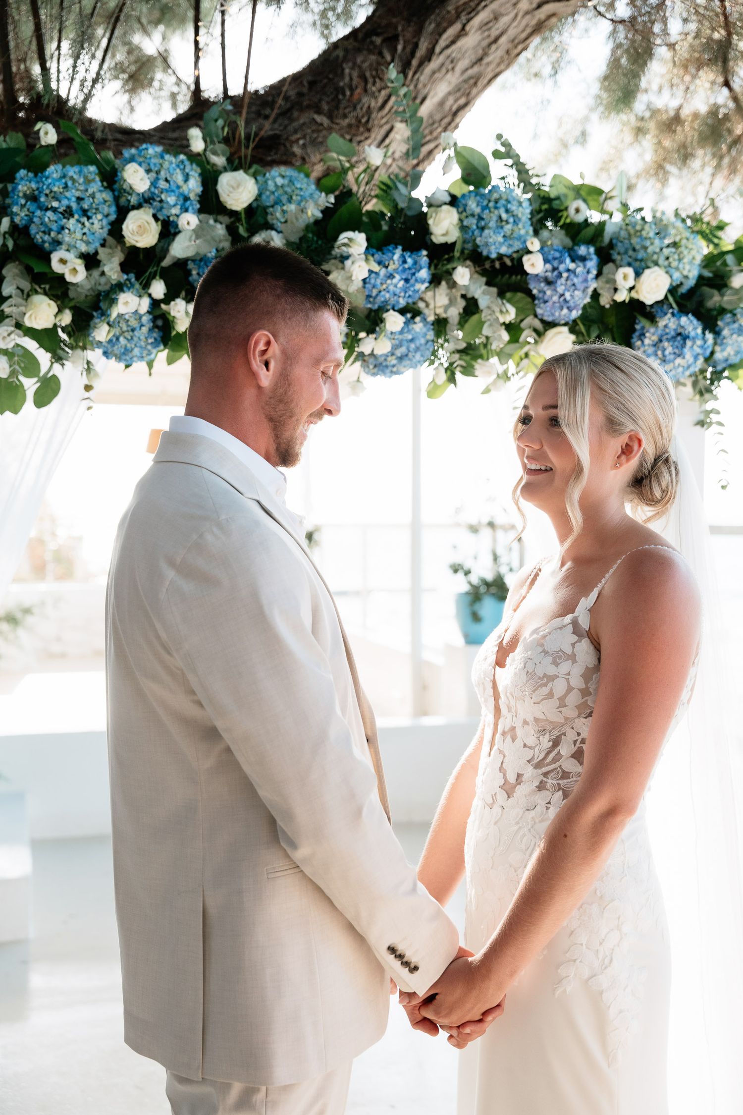 A romantic wedding scene under an arch decorated with blue hydrangeas and white flowers as a couple holds hands.