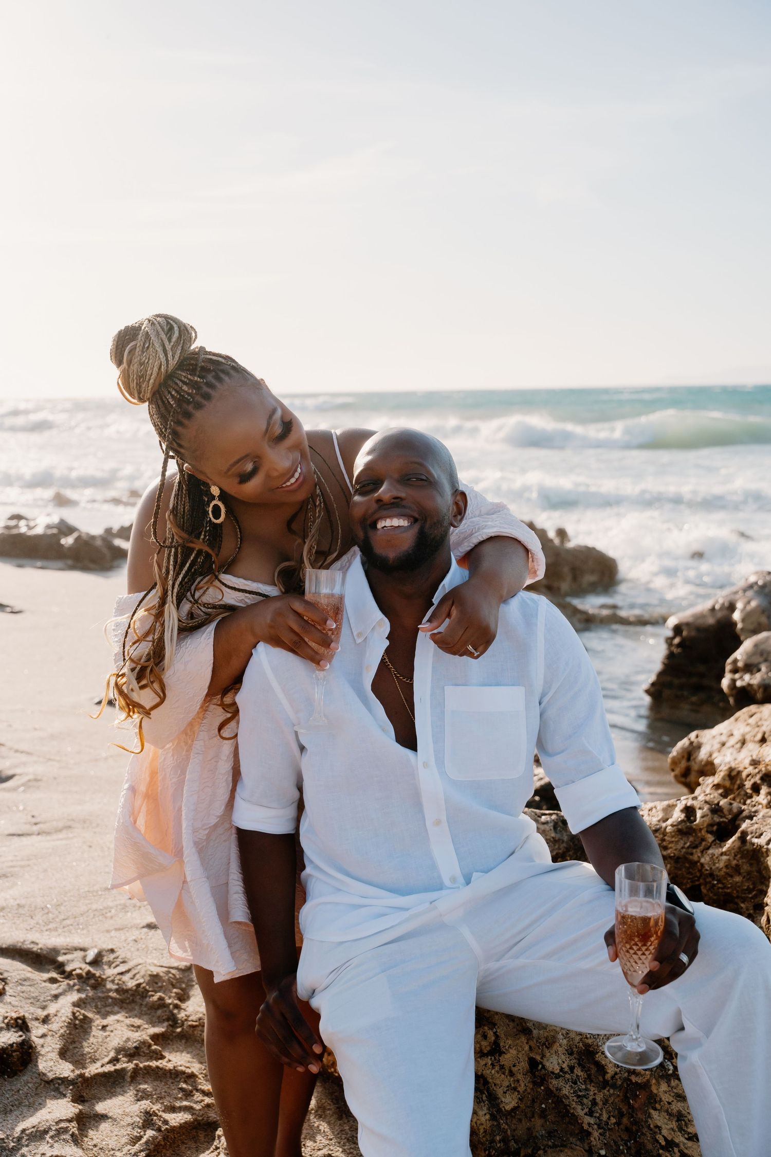 Romantic couple in white outfits share intimate moments together on a rocky beach at sunset.