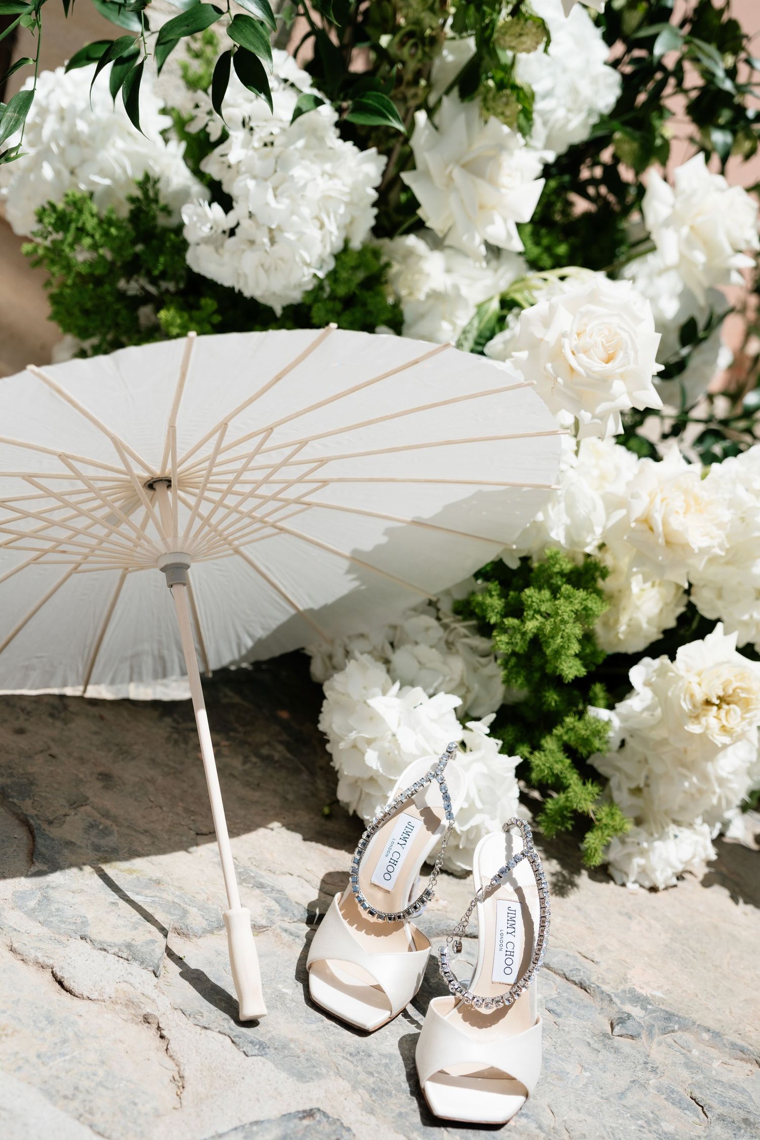 White Jimmy Choo bridal heels beside a parasol umbrella, surrounded by lush white peonies and roses on stone steps.