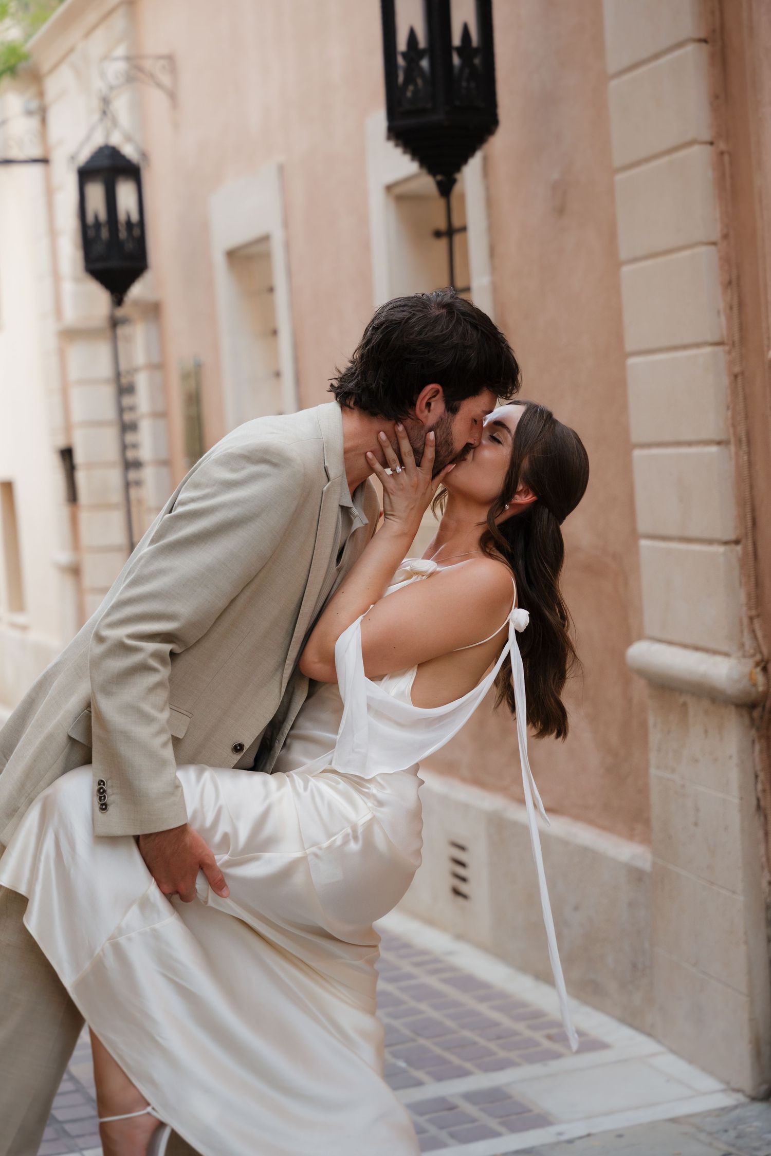 A romantic couple embraces and kisses on a Mediterranean style street with lanterns and peach colored walls.