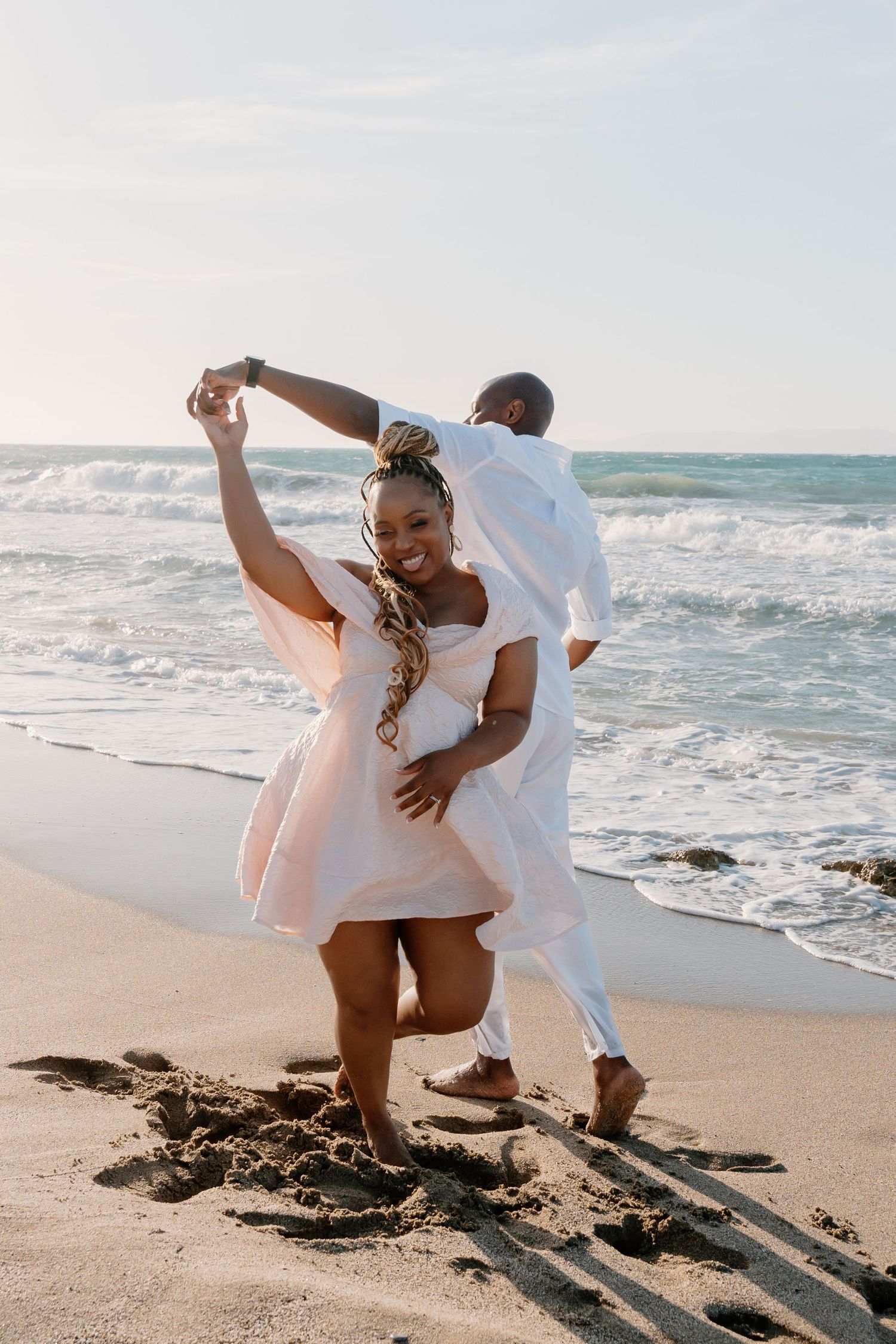 A couple in white outfits dances joyfully together on a beach with ocean waves in the background.