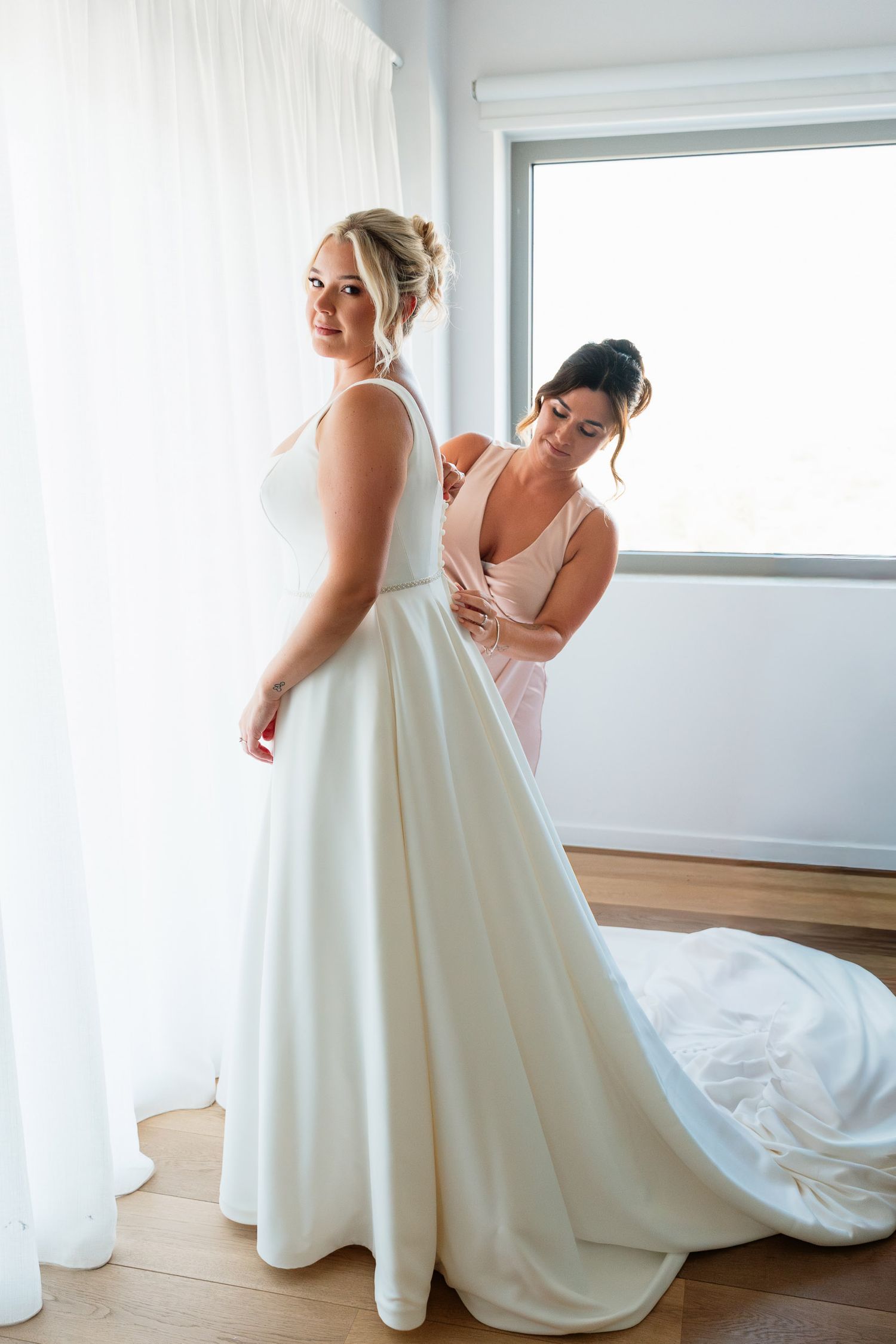 Bride in elegant white ball gown getting ready in bright sunlit room while bridesmaids assist with final touches.