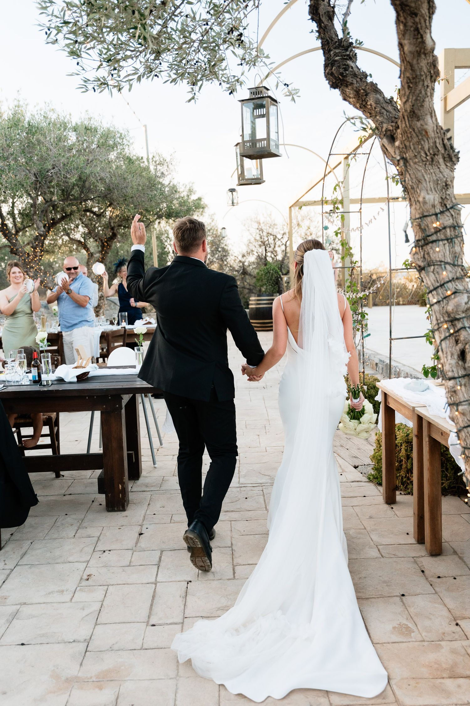 A wedding couple walks along a Mediterranean stone terrace with olive trees and lanterns creating a romantic atmosphere.
