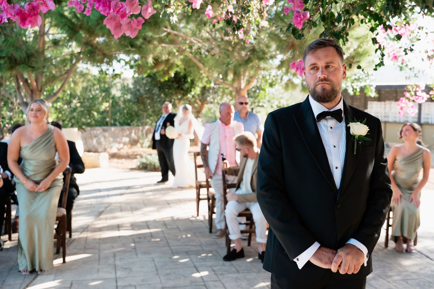Wedding party members walk down an outdoor pathway lined with pink flowers and greenery during a summer celebration.