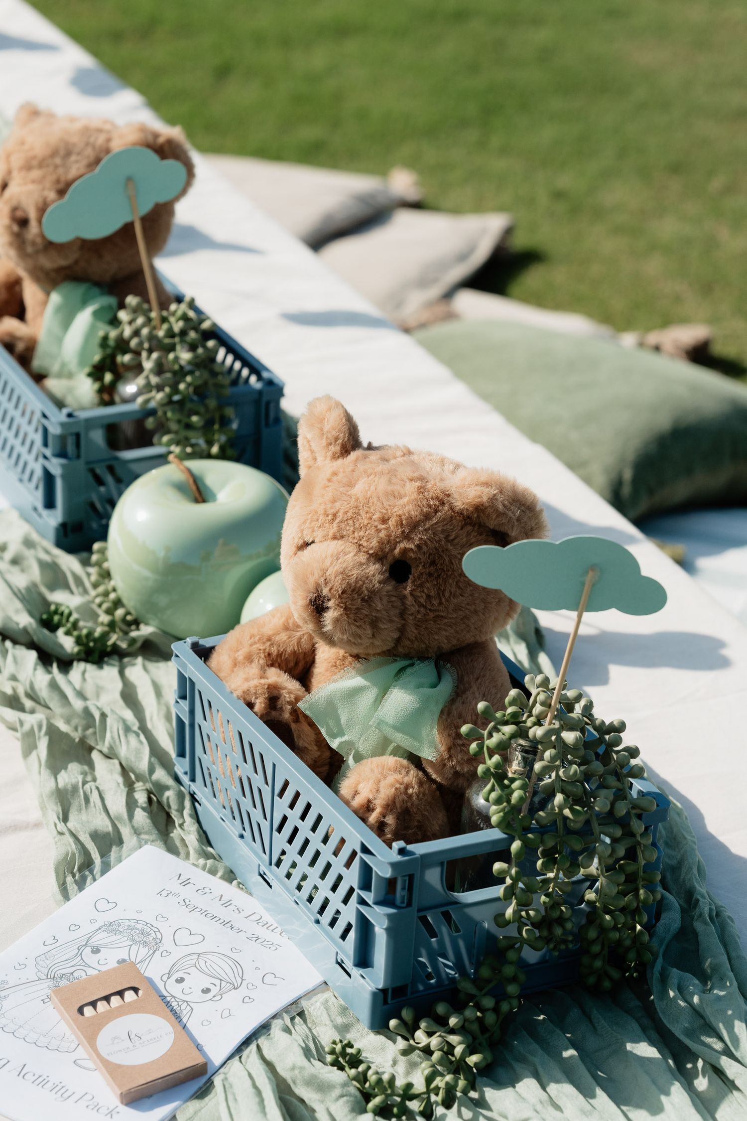 Teddy bear decorations with greenery and blue accents arranged on a white surface in a rustic outdoor setting.
