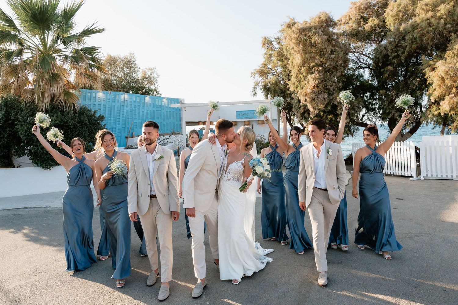 Wedding party in blue and beige attire poses together on a sunny beach boardwalk at sunset.