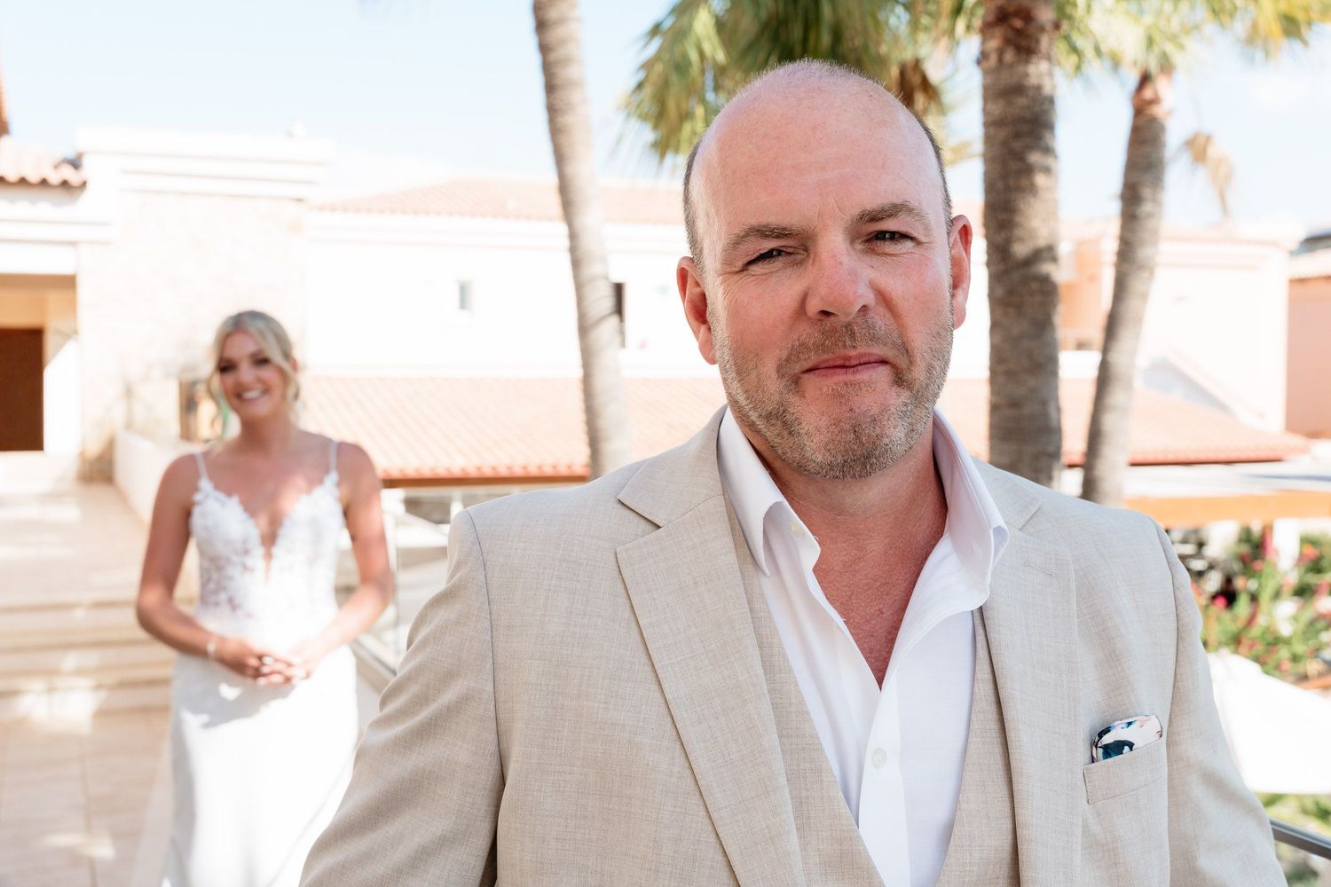 A person in a light beige suit and white shirt stands in a sunny courtyard with palm trees and white buildings.