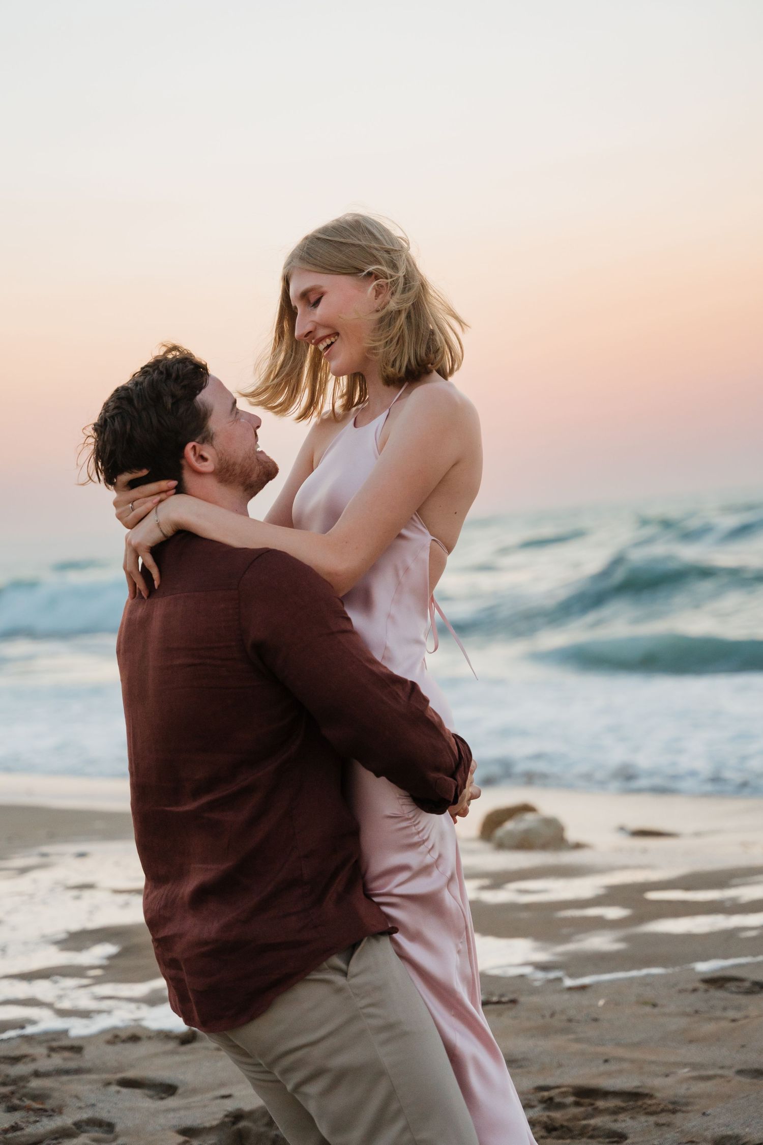 A couple embraces romantically on a beach at sunset with ocean waves in the background.