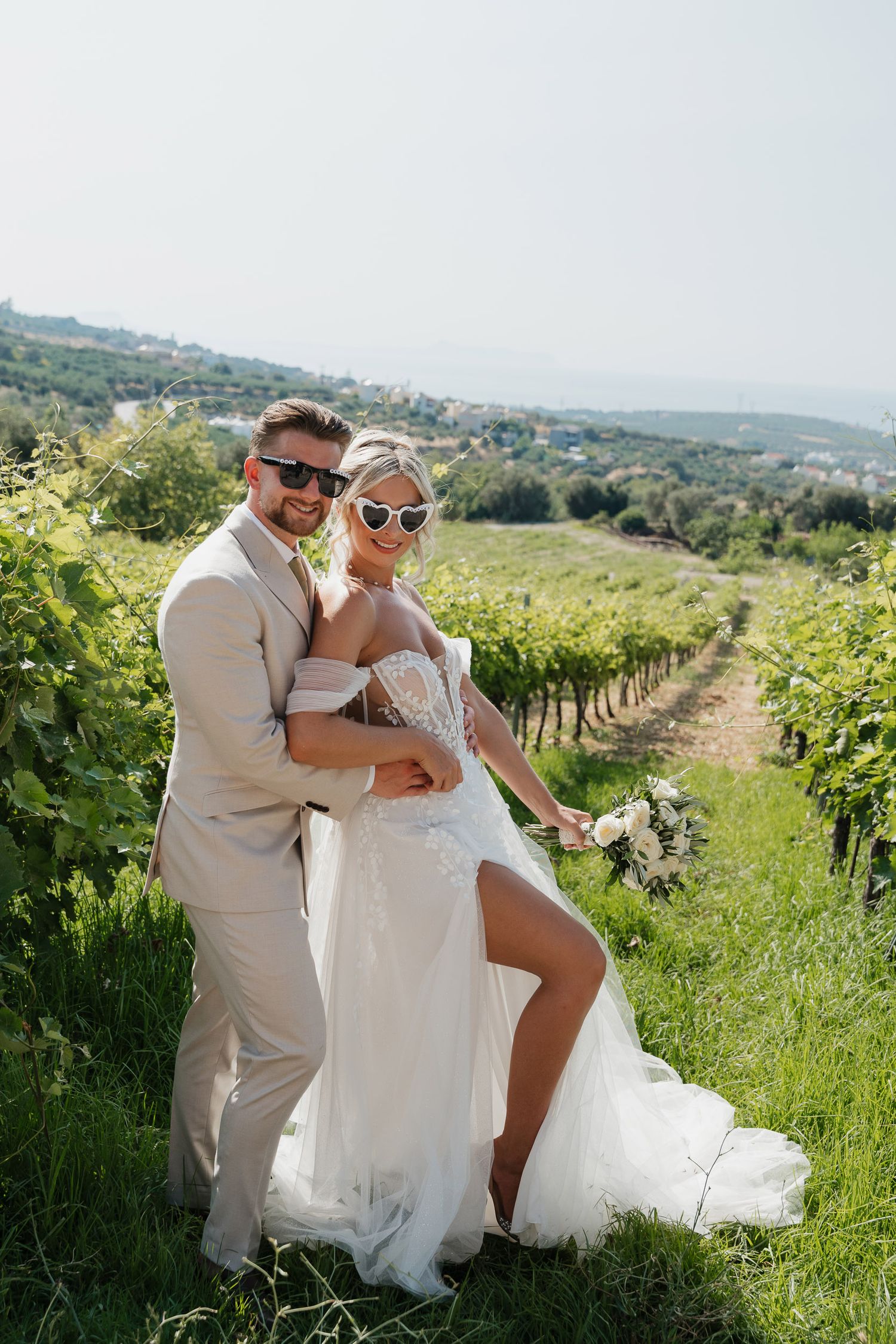 A couple poses together in a scenic vineyard on a sunny day, surrounded by rows of grapevines and rolling hills.