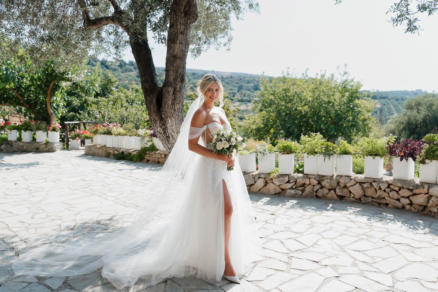 A bride in a flowing white gown with a thigh-high slit holds a bouquet in a sunny Mediterranean garden setting.