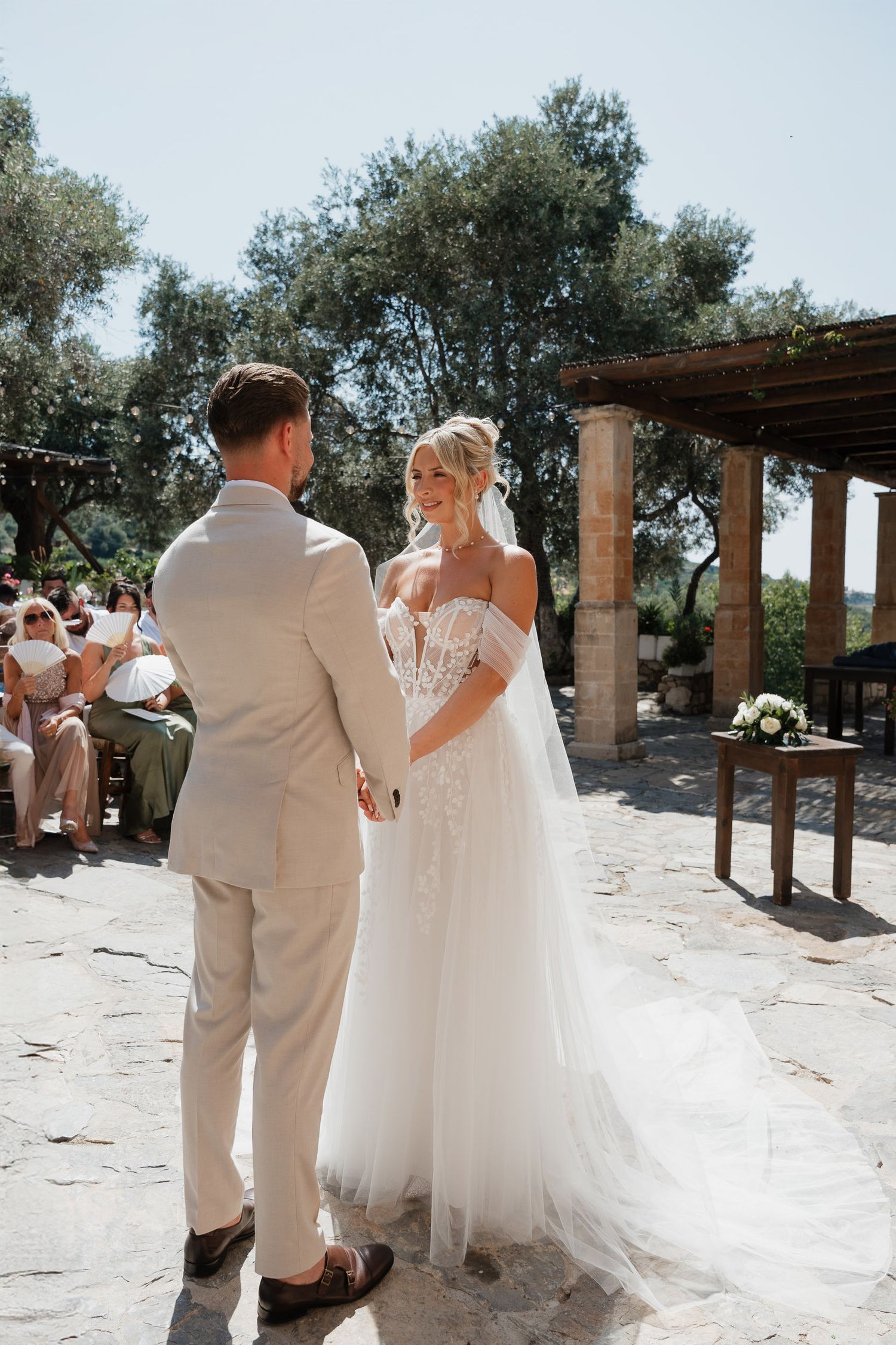 A romantic outdoor wedding ceremony takes place under a rustic wooden pergola surrounded by olive trees on a sunny day.